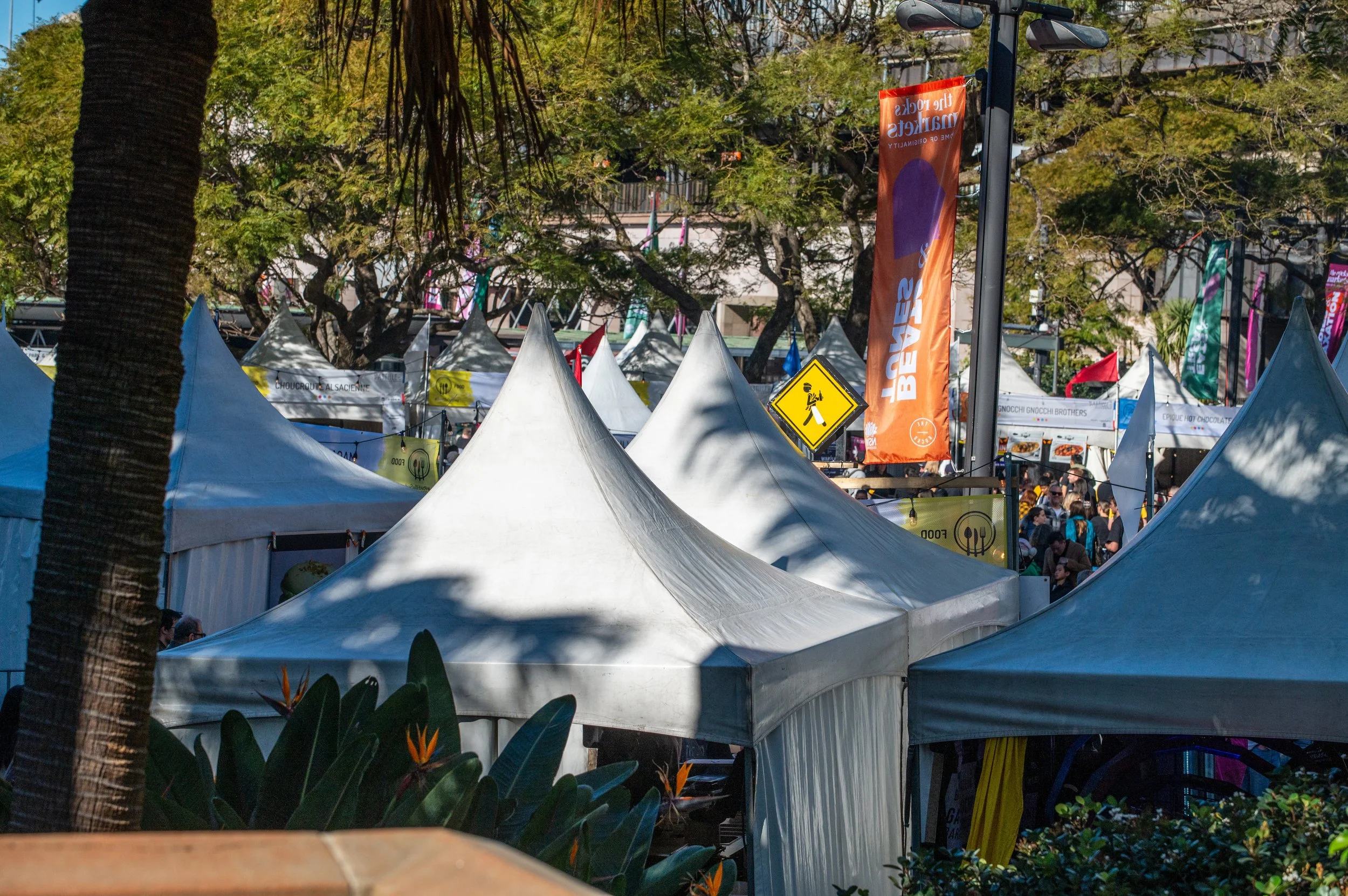 Outdoor market with white tents, colorful banners, and a large yellow pedestrian crossing sign, surrounded by trees and plants.