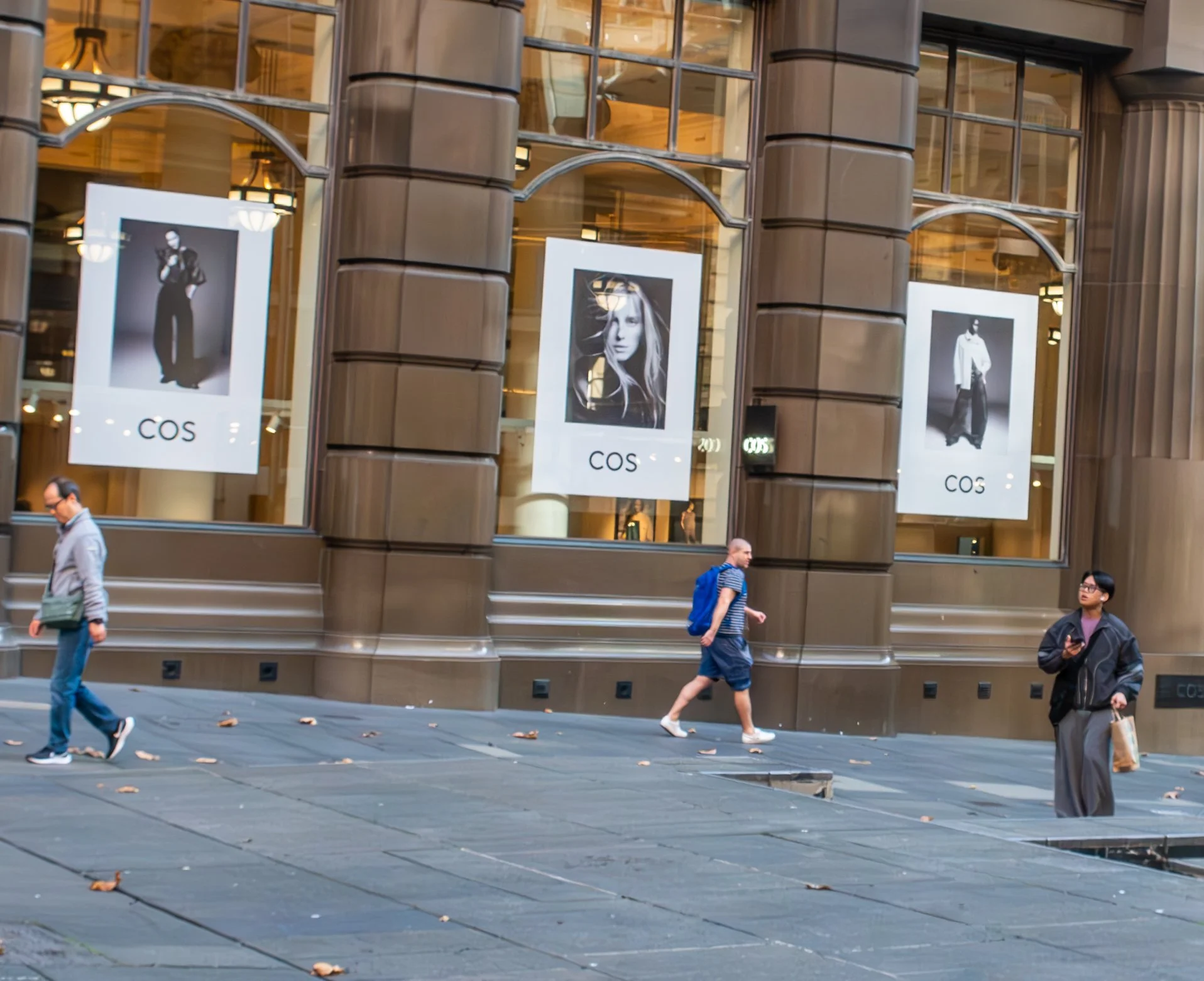 Street scene with three pedestrians walking past a large store with tall glass windows displaying fashion advertisements. The store showcases black-and-white photos of models with the brand name CO$ underneath each image.