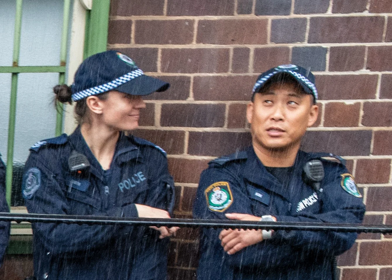 Two police officers standing together in rain against a brick wall, smiling and looking at each other.