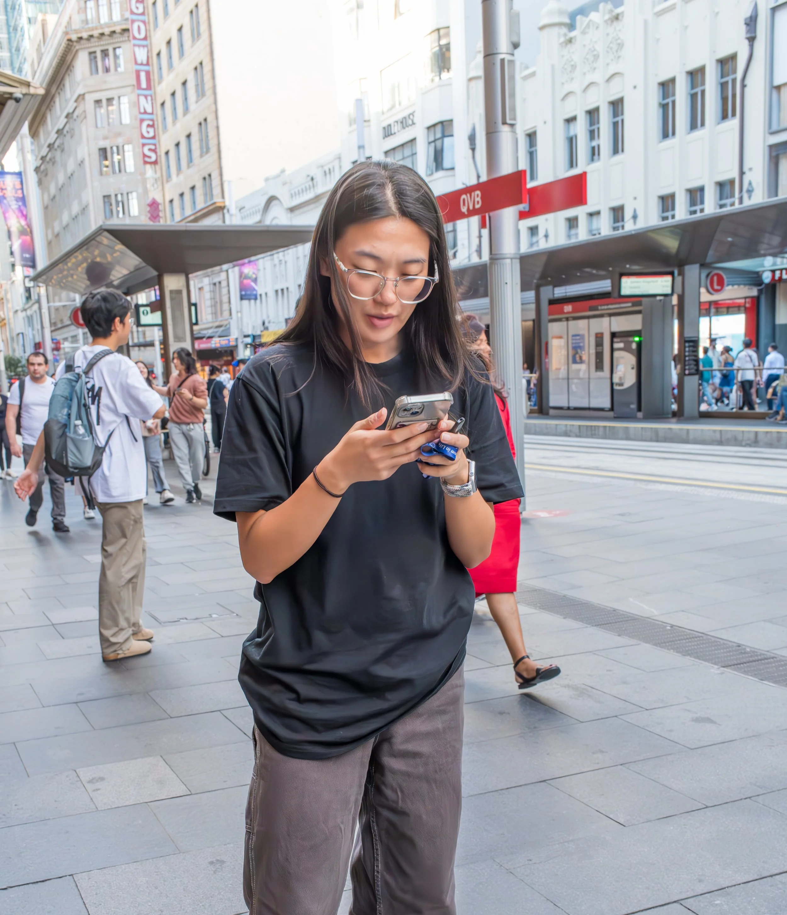 A young woman in glasses and a black T-shirt looking at her phone while walking on a busy city sidewalk.
