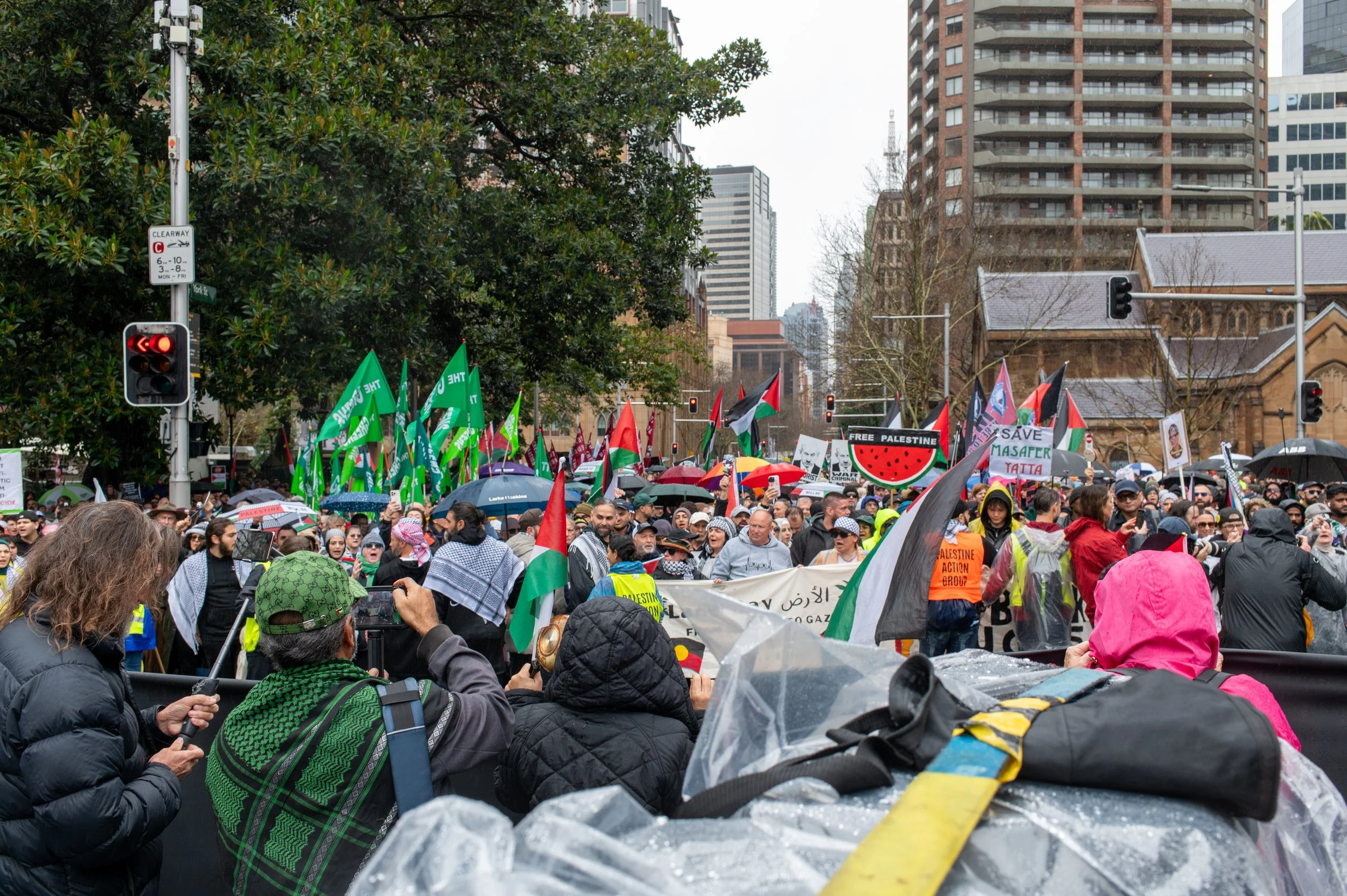 A large crowd of people gathered in a city street, holding various protest signs and flags, some with messages supporting Palestine, under gray weather with some umbrellas.