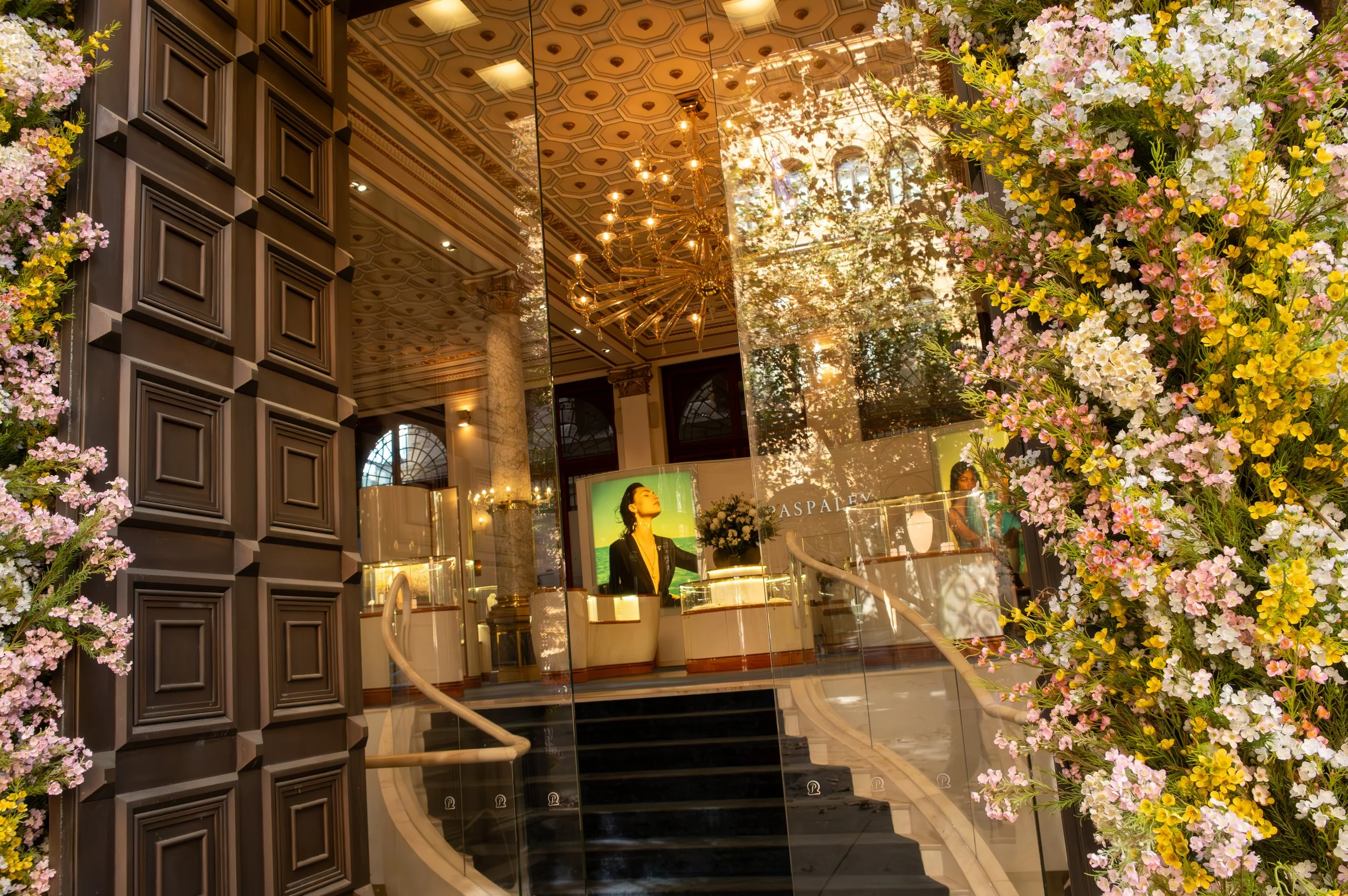 Interior view of a luxurious jewelry store entrance with a staircase, glass walls, floral decorations, and a digital advertisement featuring a woman with short dark hair.