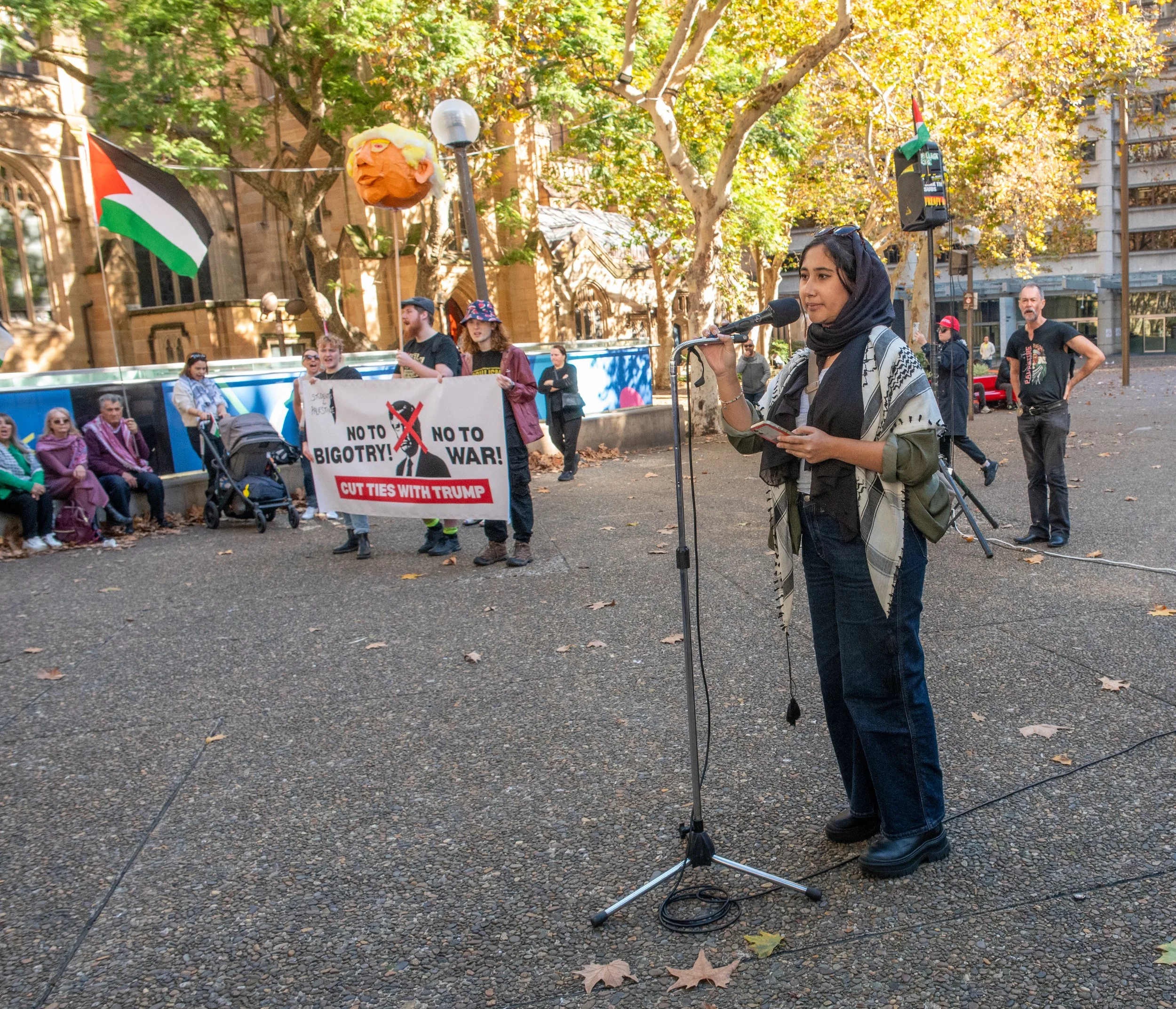 A woman in a headscarf speaking into a microphone at a protest, with a group holding a banner that reads 'No to Bigotry! No to War! Cut Ties with Trump' and a Palestinian flag in the background, outdoors on a city street surrounded by trees.