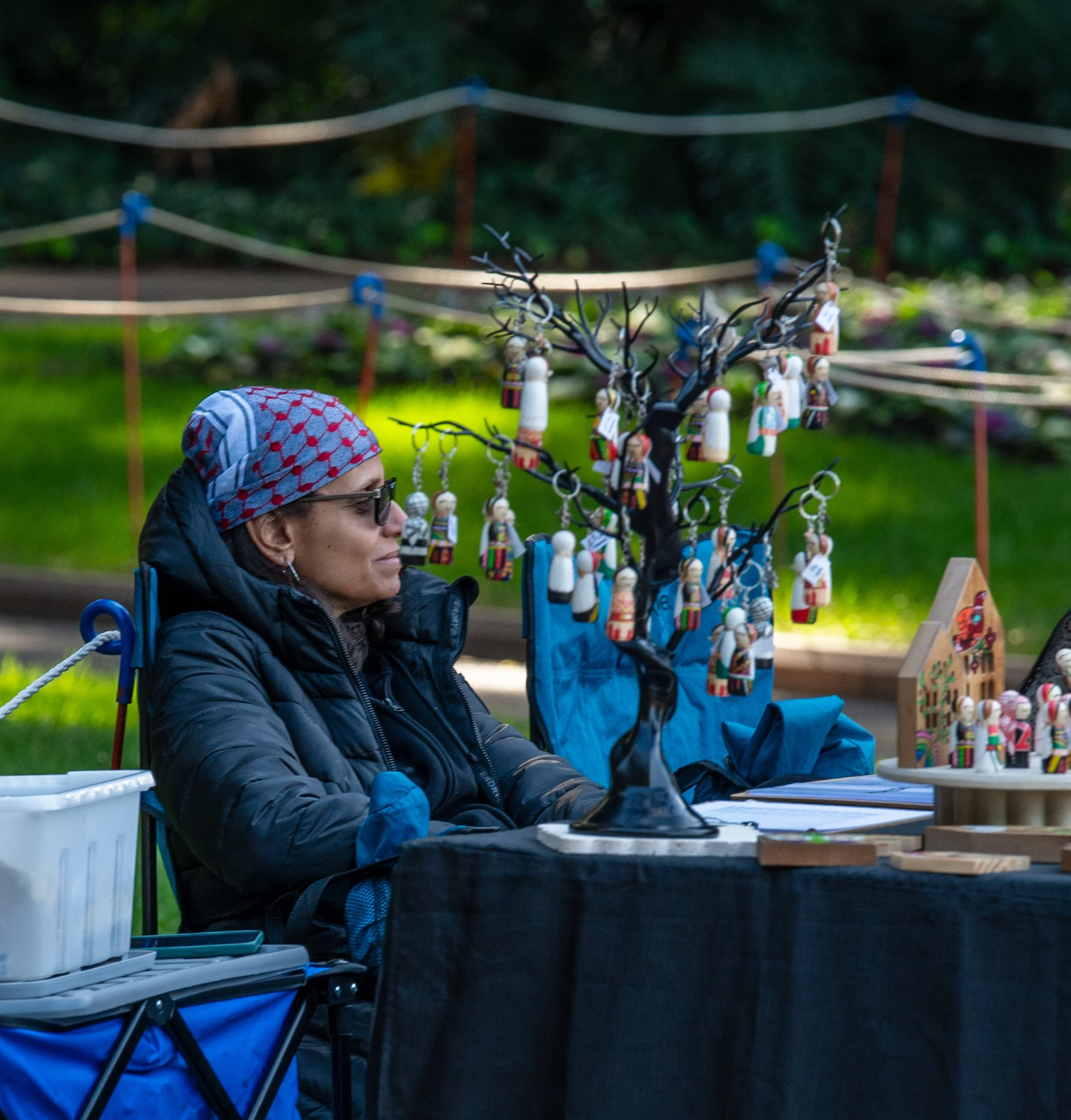 A woman wearing a patterned headscarf, sunglasses, and a dark jacket sits at a table displaying handcrafted wooden figurines and miniature trees decorated with similar figures, outdoors with a blurred background of greenery and a decorative rope fencing.