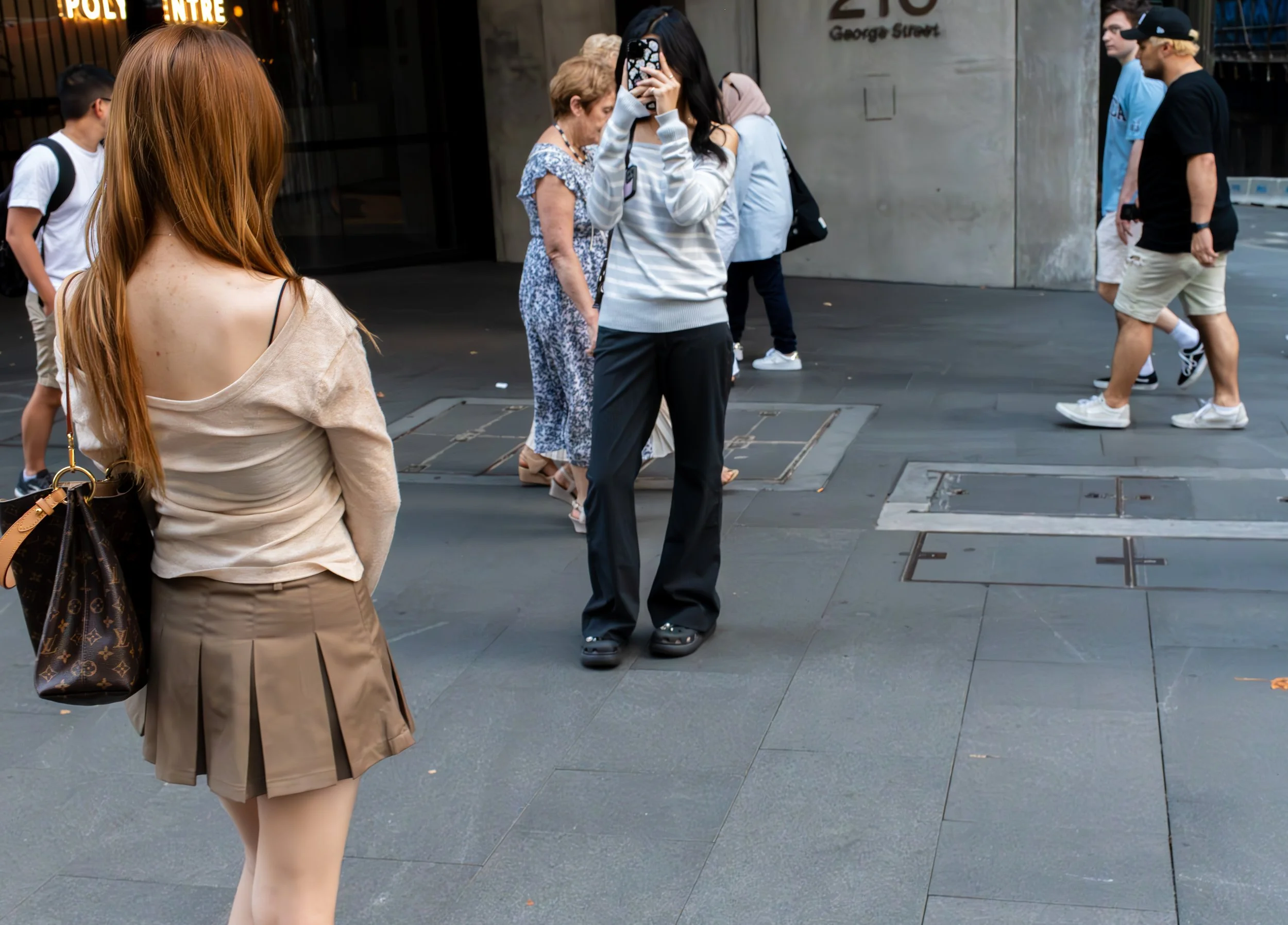 A woman in a beige off-the-shoulder top and a pleated skirt with a Louis Vuitton bag, standing on a city sidewalk, facing a woman with black hair using her phone. Several other pedestrians walk nearby, and a building with a sign reading 'George Street' is in the background.