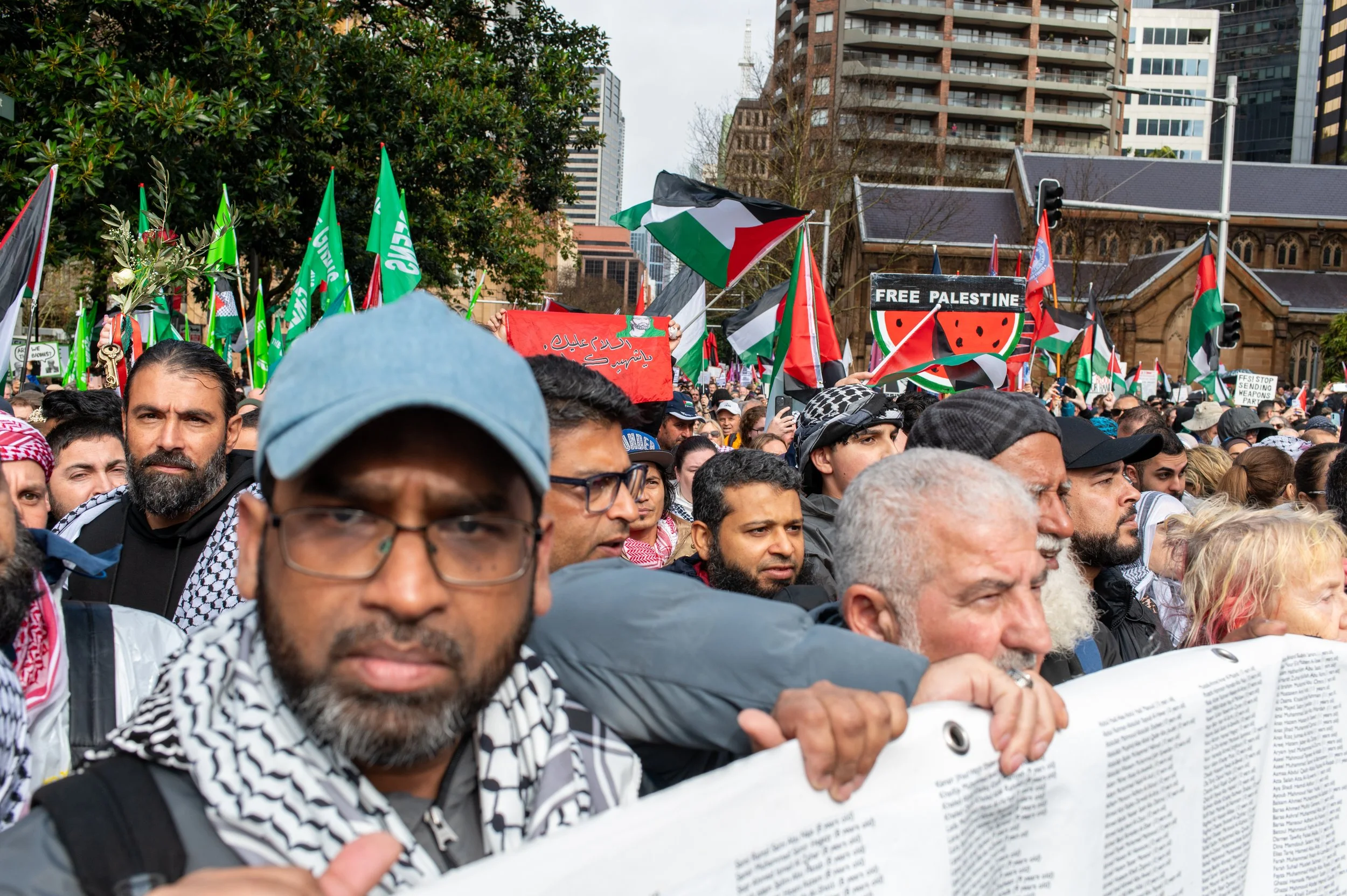Crowd of protesters holding signs and flags, with some signs reading "Free Palestine" and others featuring green, black, red, and white colors, in an urban setting.