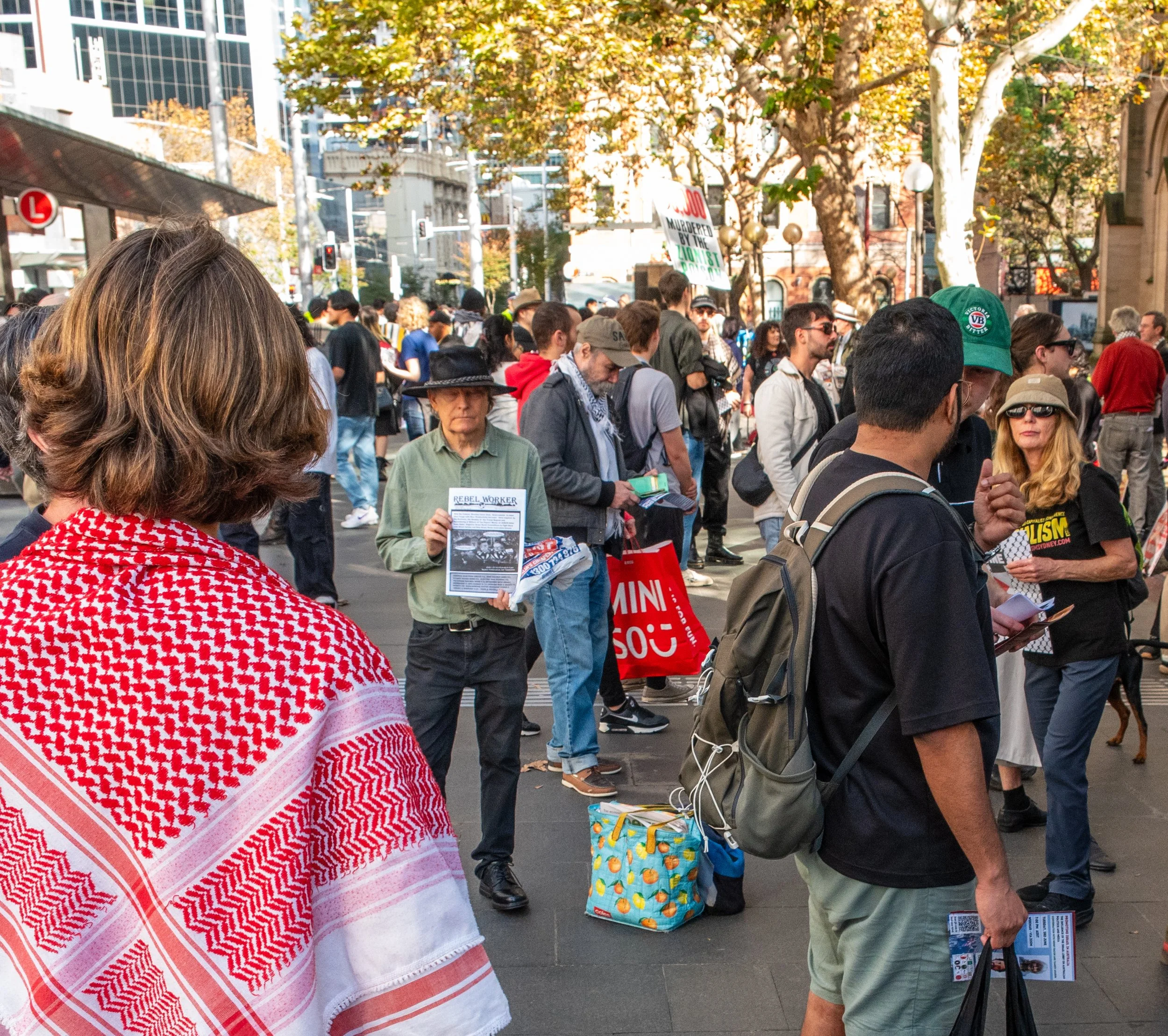 Crowd of people gathered outdoors in a city square, some holding signs and flyers, trees with autumn-colored leaves in the background.