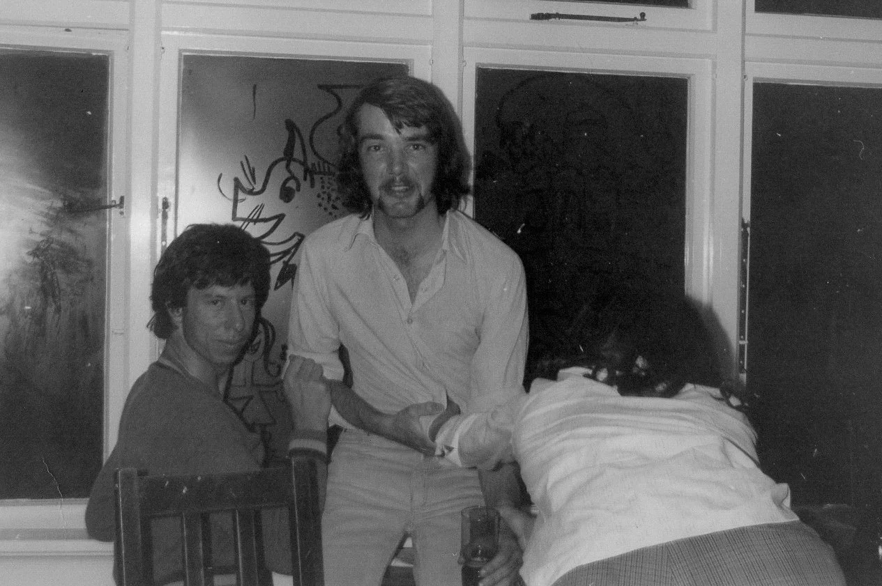 Three people in a black-and-white photo inside a room with artwork behind them. One person is seated at a table, another is standing, and the third has their back to the camera.