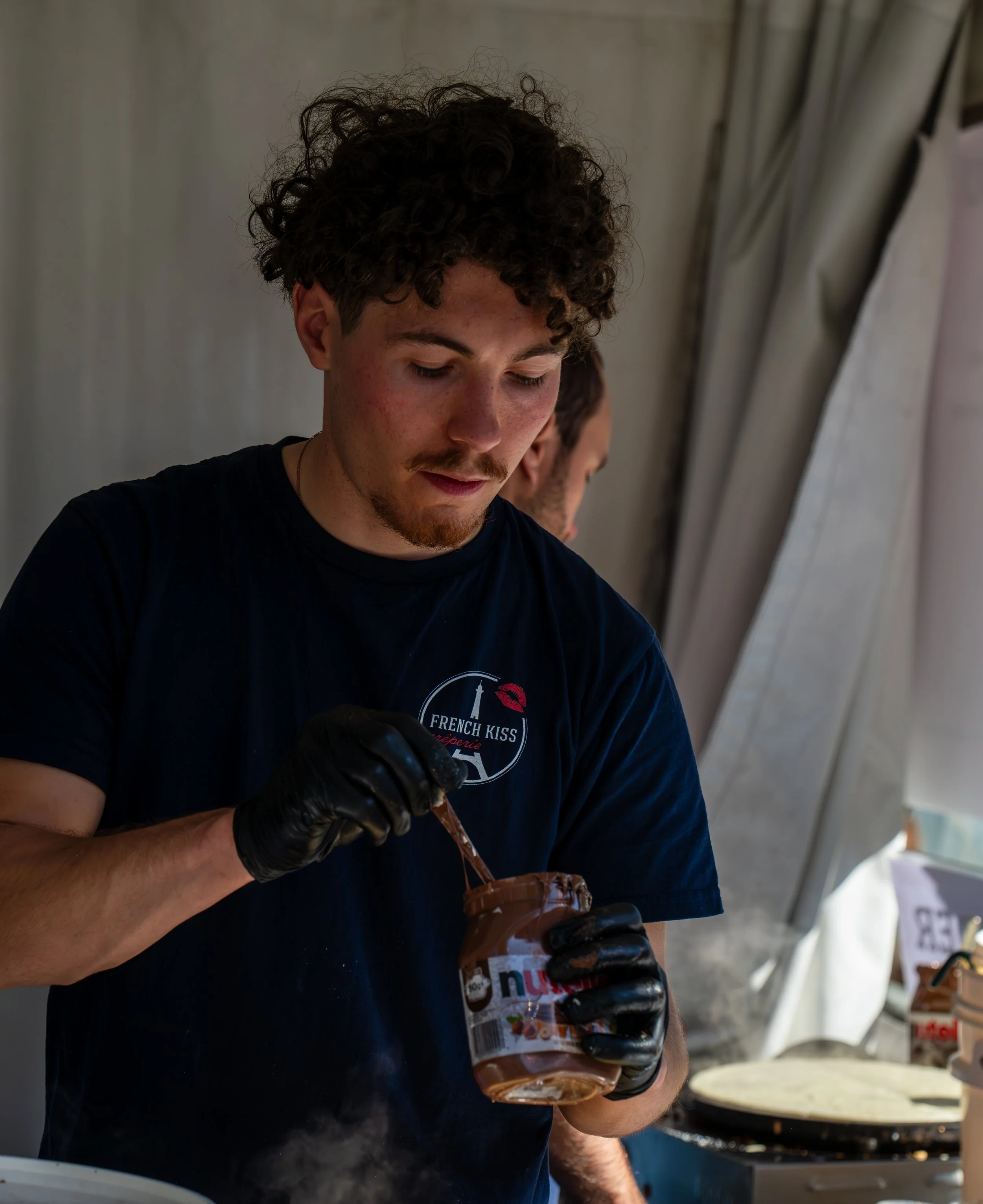 Young man with curly hair wearing a black t-shirt and black gloves spreading Nutella on a piece of bread.