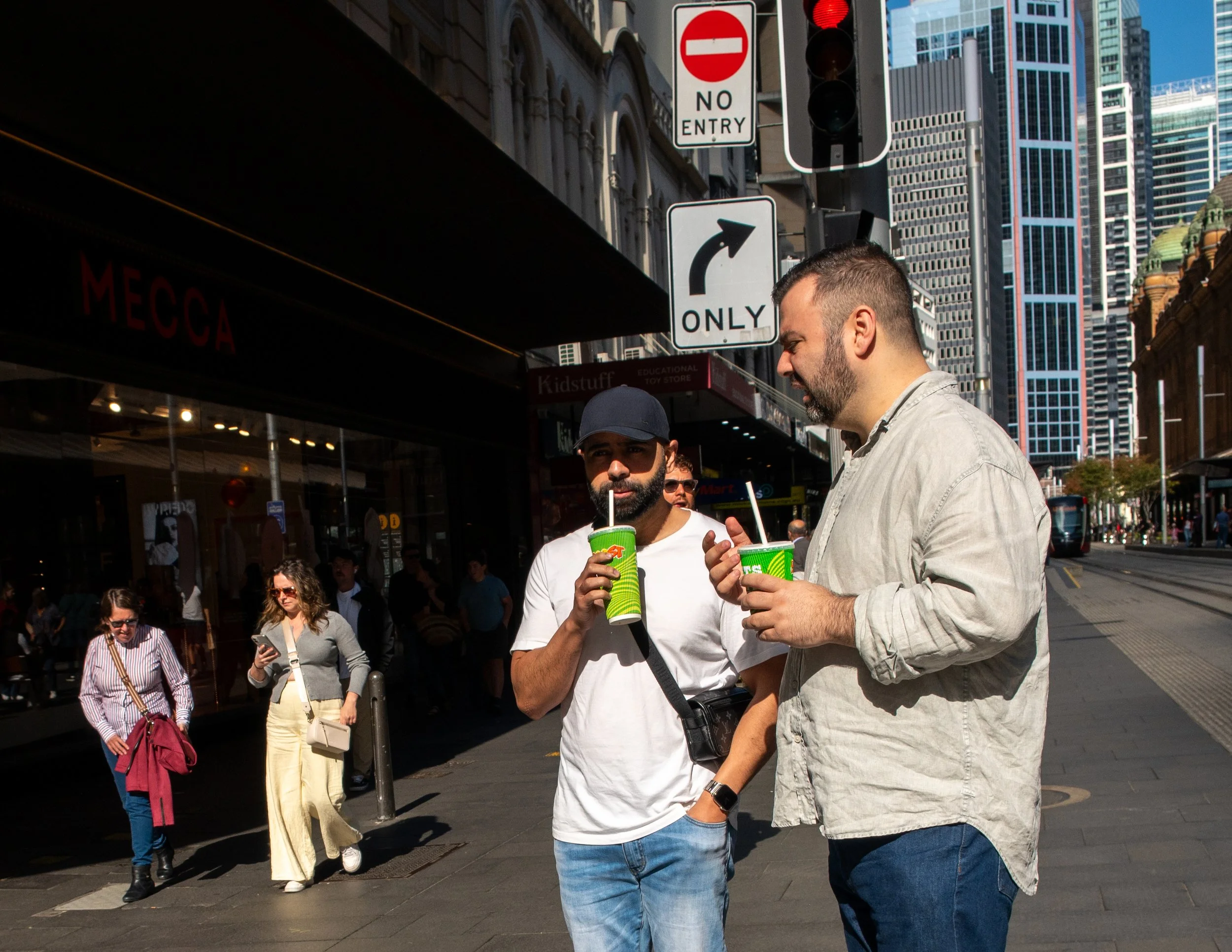 Two men stand on a city street holding green cups with straws, engaged in conversation, with pedestrians and tall buildings in the background.