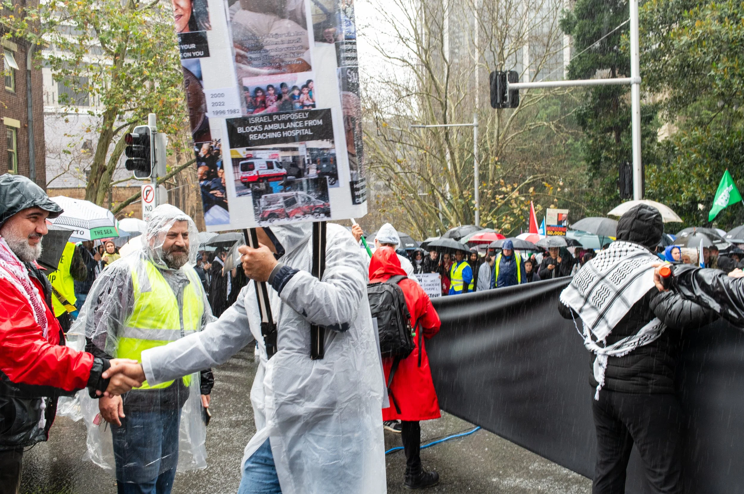 People protesting in the rain, some wearing raincoats and holding umbrellas, with signs and banners. Two individuals are shaking hands, and others are standing or marching behind a large black banner.