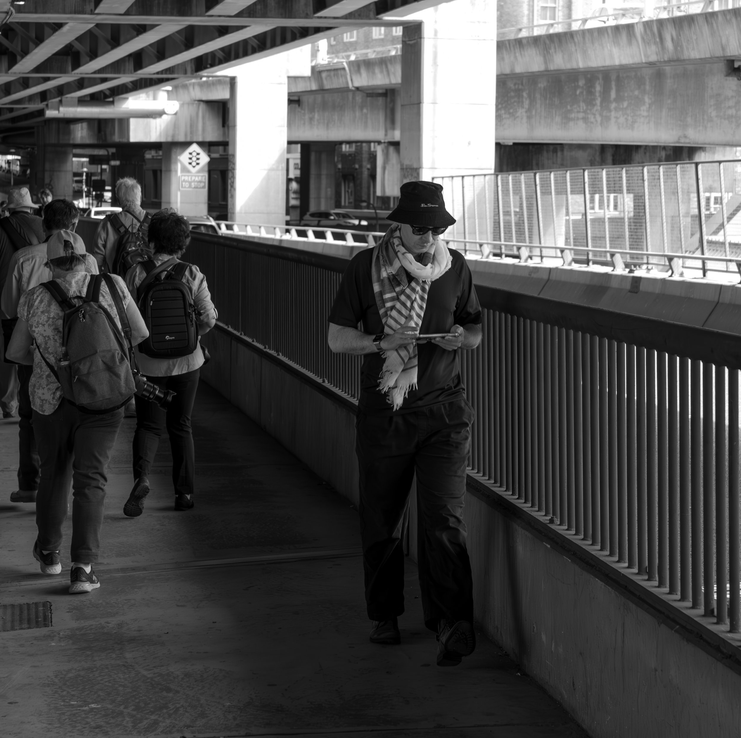 A person wearing a hat, sunglasses, and a scarf walks along a sidewalk while looking at their phone, with other people nearby under an overpass.