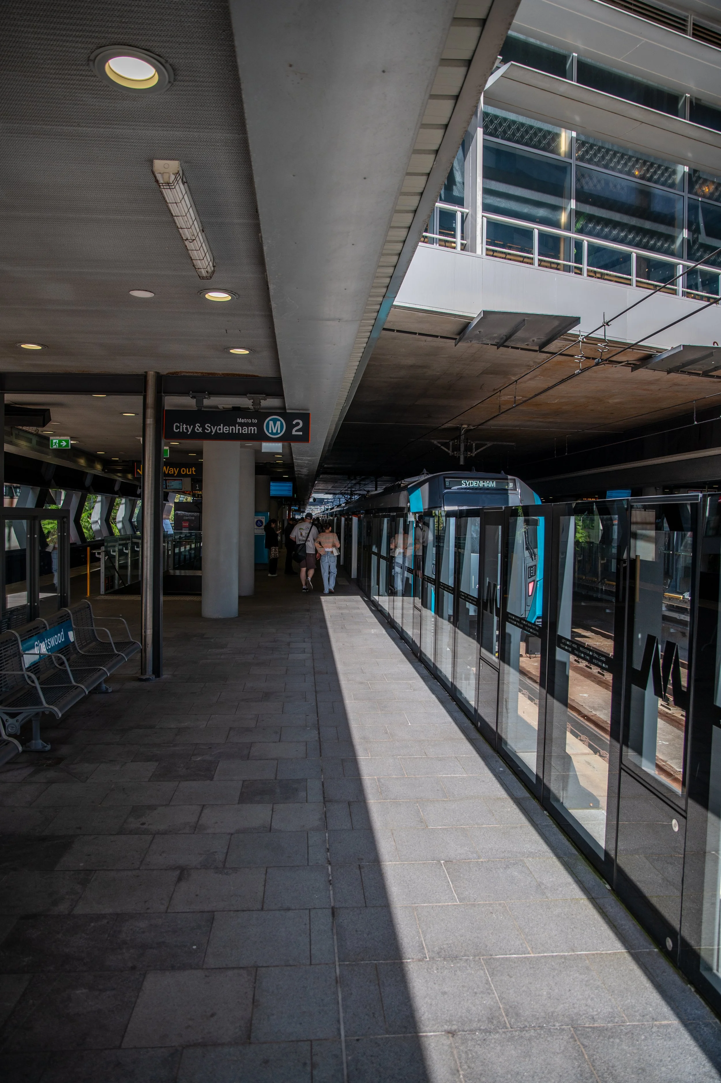 Platform at a modern train station with a blue train and passengers waiting, signage indicating Metro Line 2 to City & Sydenham, and benches on the left side.