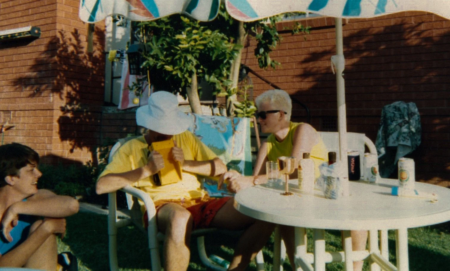 Three people enjoying a summer gathering outdoors around a white table with drinks, snacks, and bottles, under a striped umbrella, beside a brick wall and lush greenery.