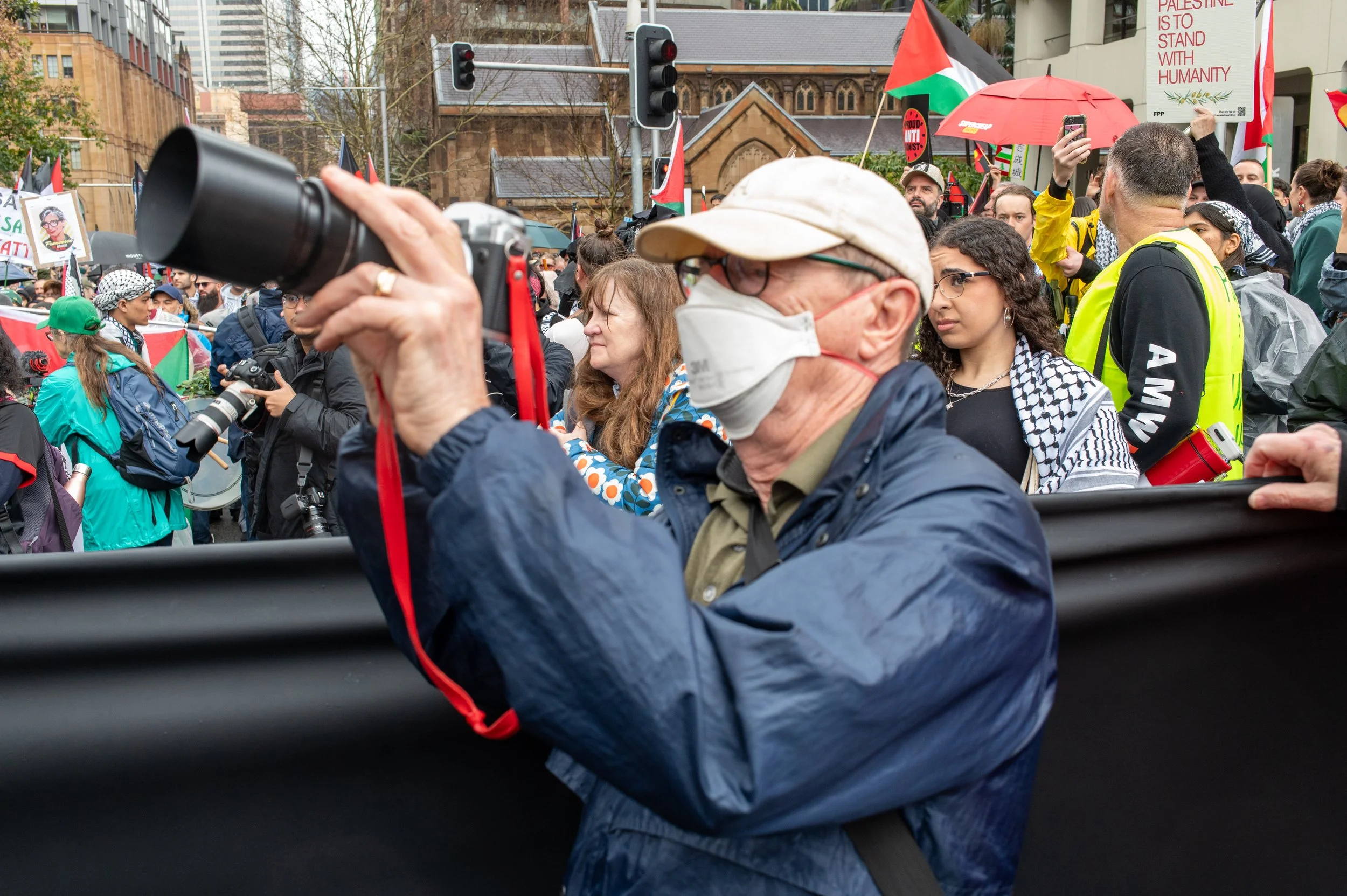 Older man wearing a face mask and glasses taking a photo with a camera at a protest or rally, with a crowd holding signs and flags in the background.