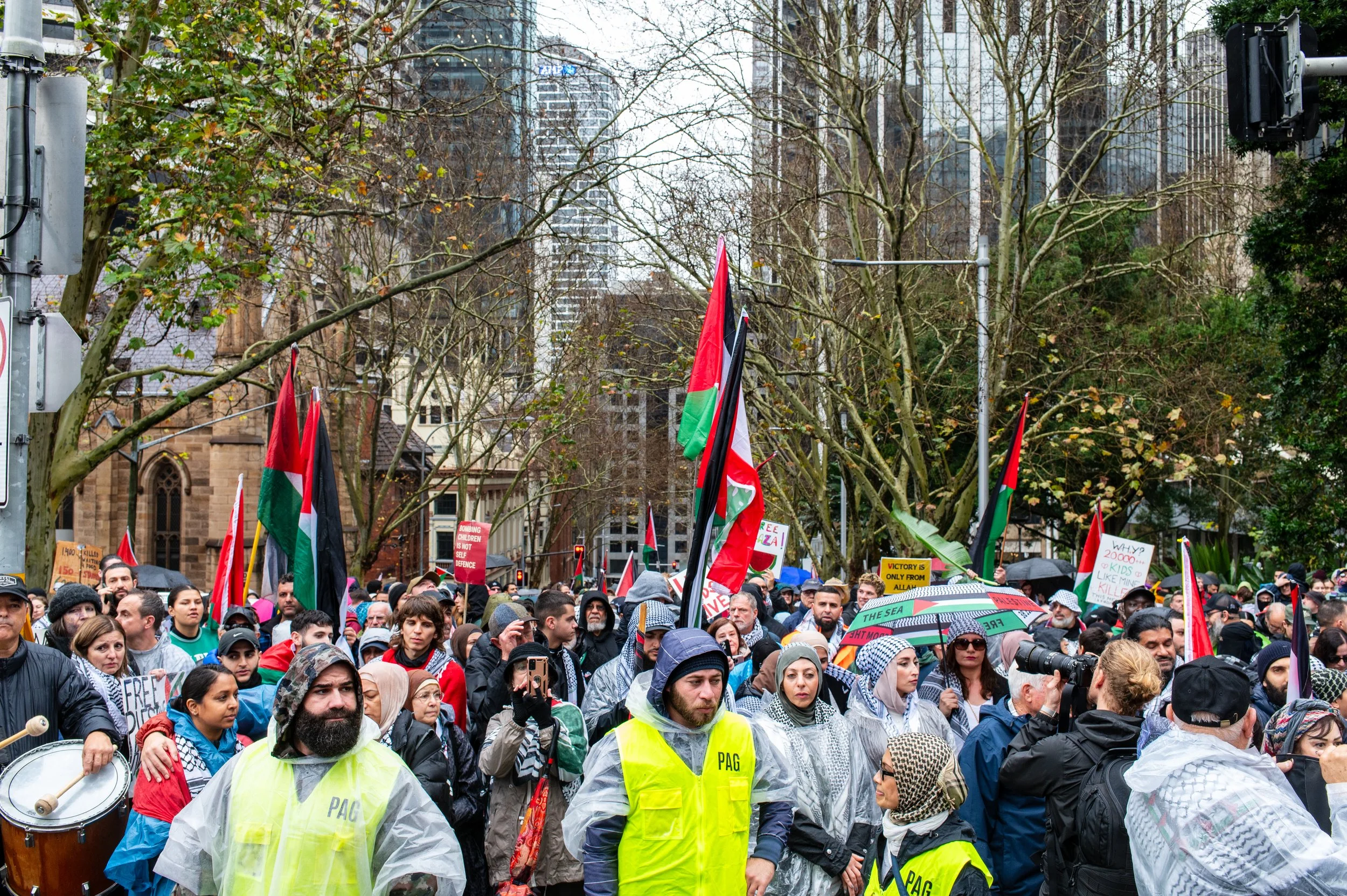 A large group of protesters gathered outdoors, holding flags, signs, and umbrellas, with some wearing raincoats. The crowd appears diverse, with some people holding signs with messages, and there are a few visible security personnel wearing yellow vests.
