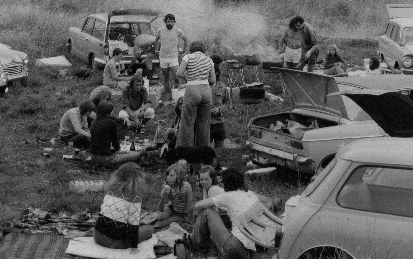 People having a picnic outdoors near parked cars on a grassy field, some sitting on blankets and others preparing food on a portable grill.
