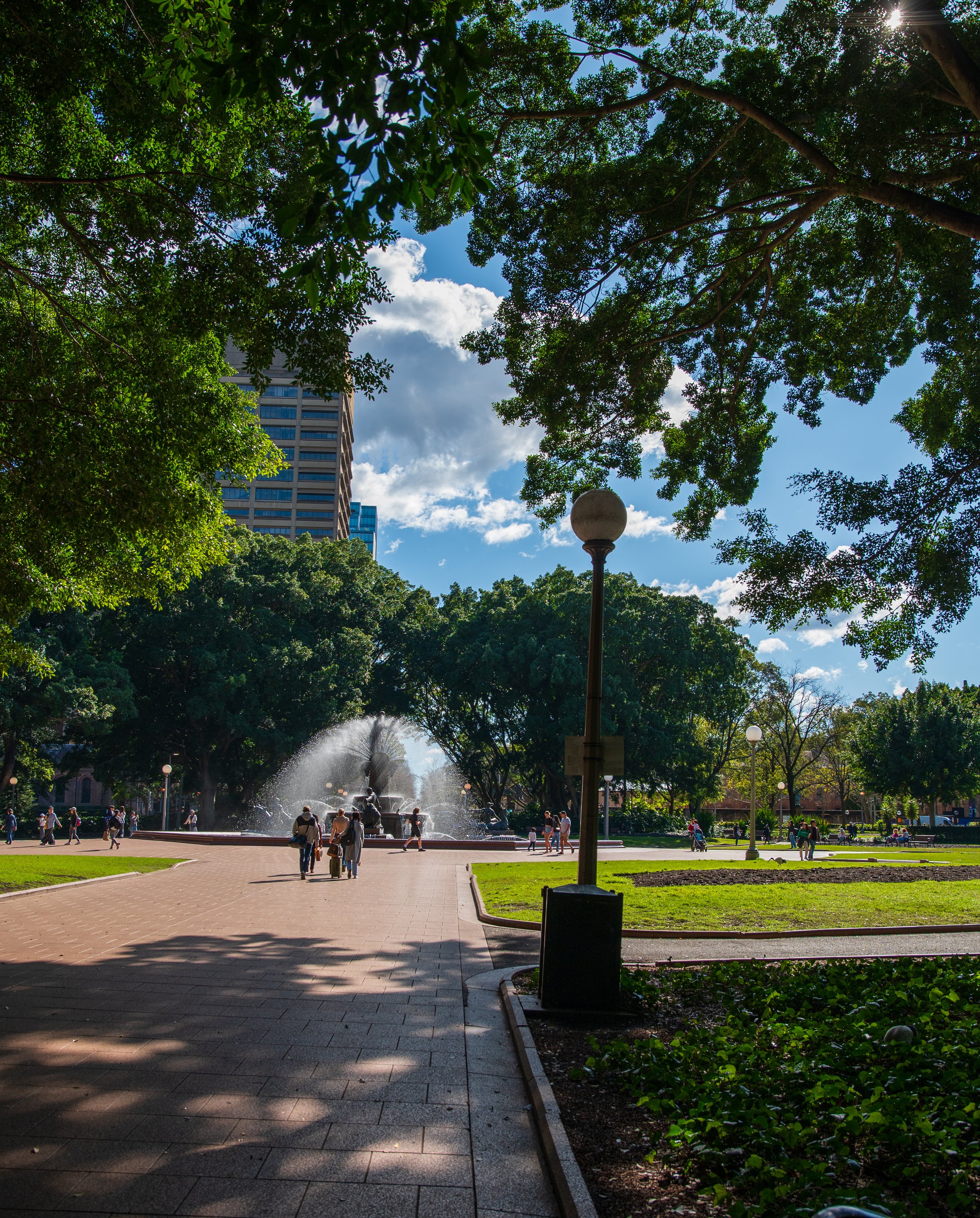 A park with a fountain and walking paths, surrounded by trees, with people strolling and tall buildings in the background under a partly cloudy sky.