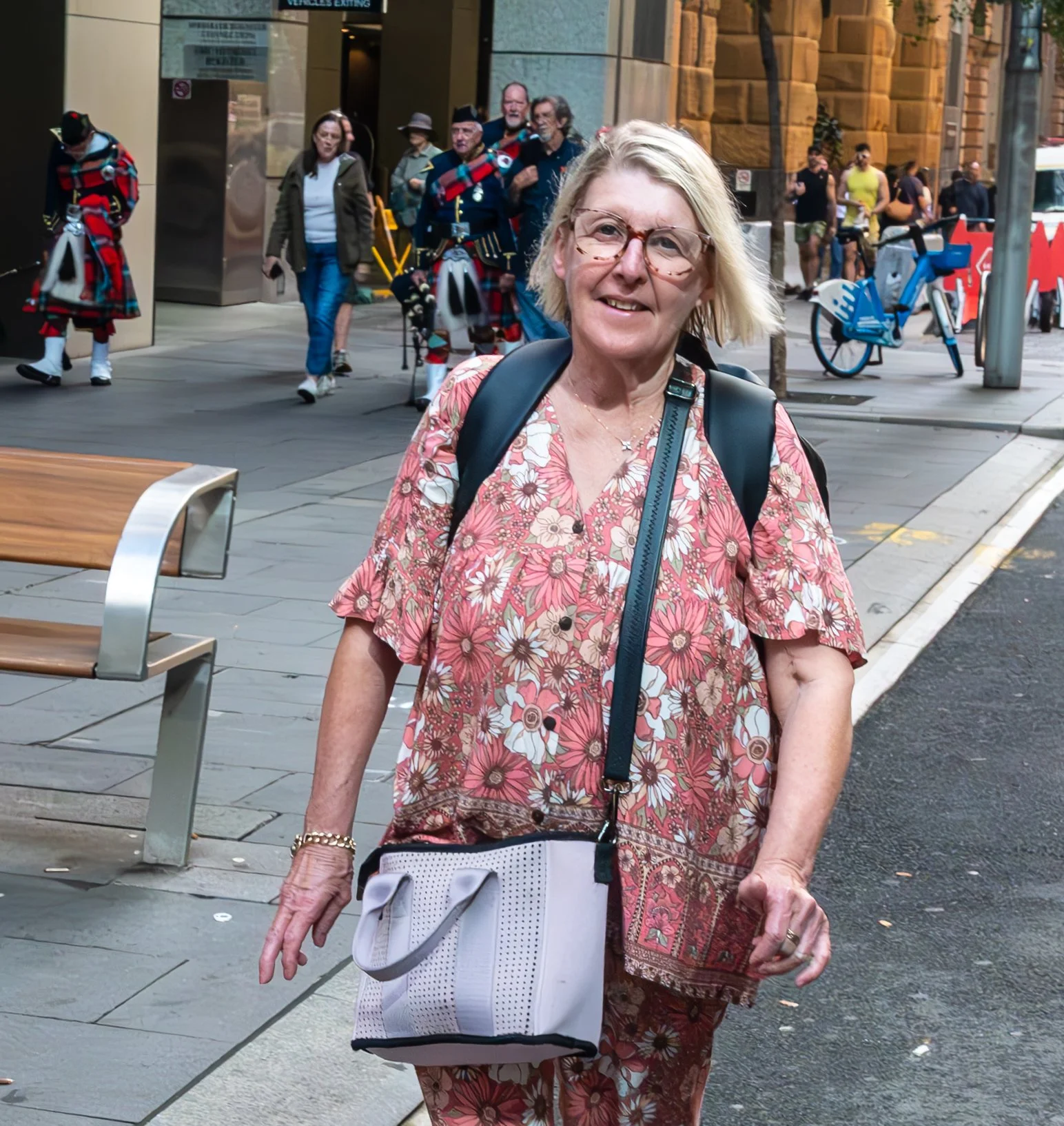 An older woman with blonde hair and glasses walking on city sidewalk, wearing a pink floral outfit, carrying a white bag, with people and performers dressed in traditional Scottish attire in the background.