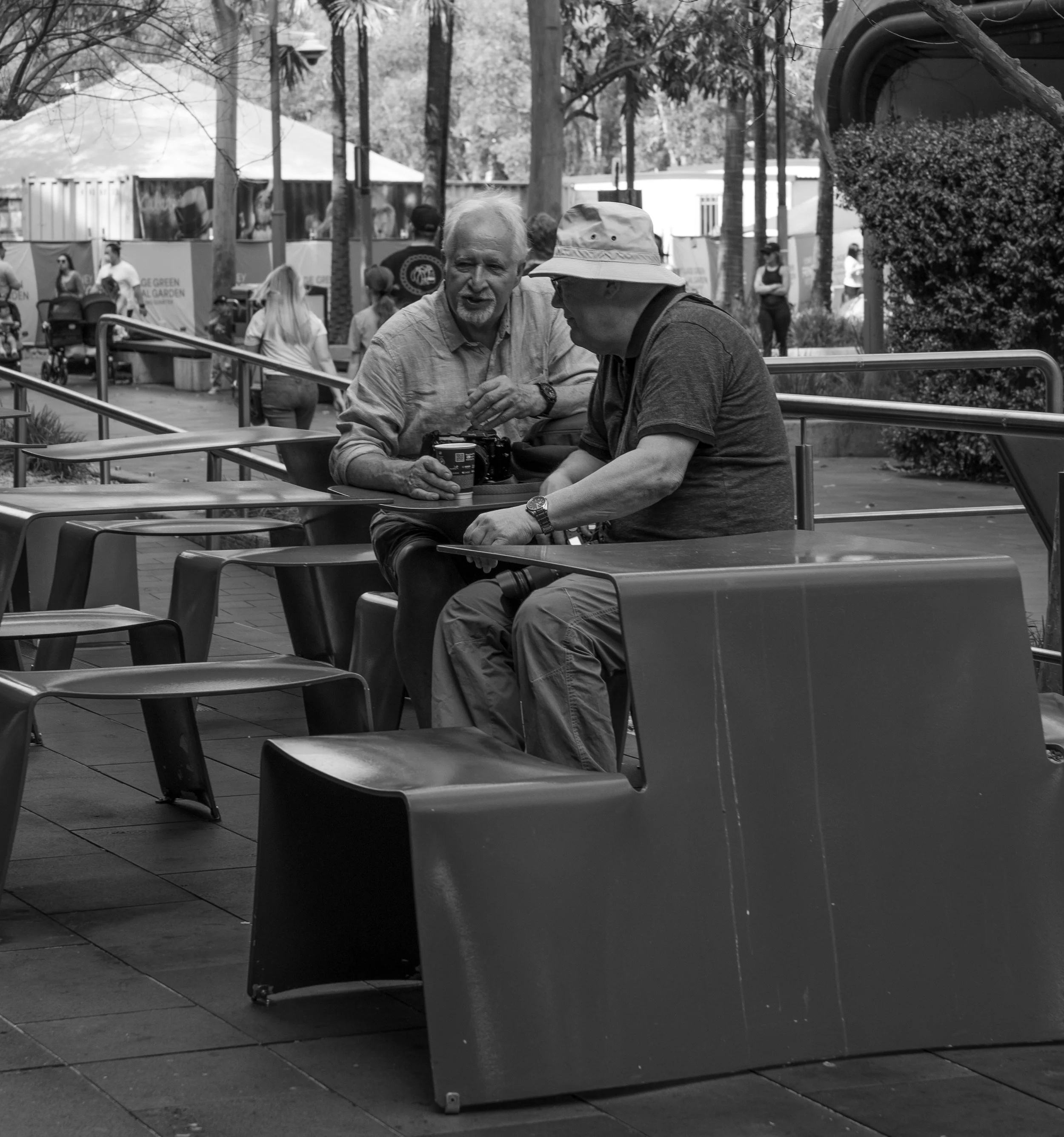 Two men sitting at a table outdoors, looking at a camera, with people walking in the background.