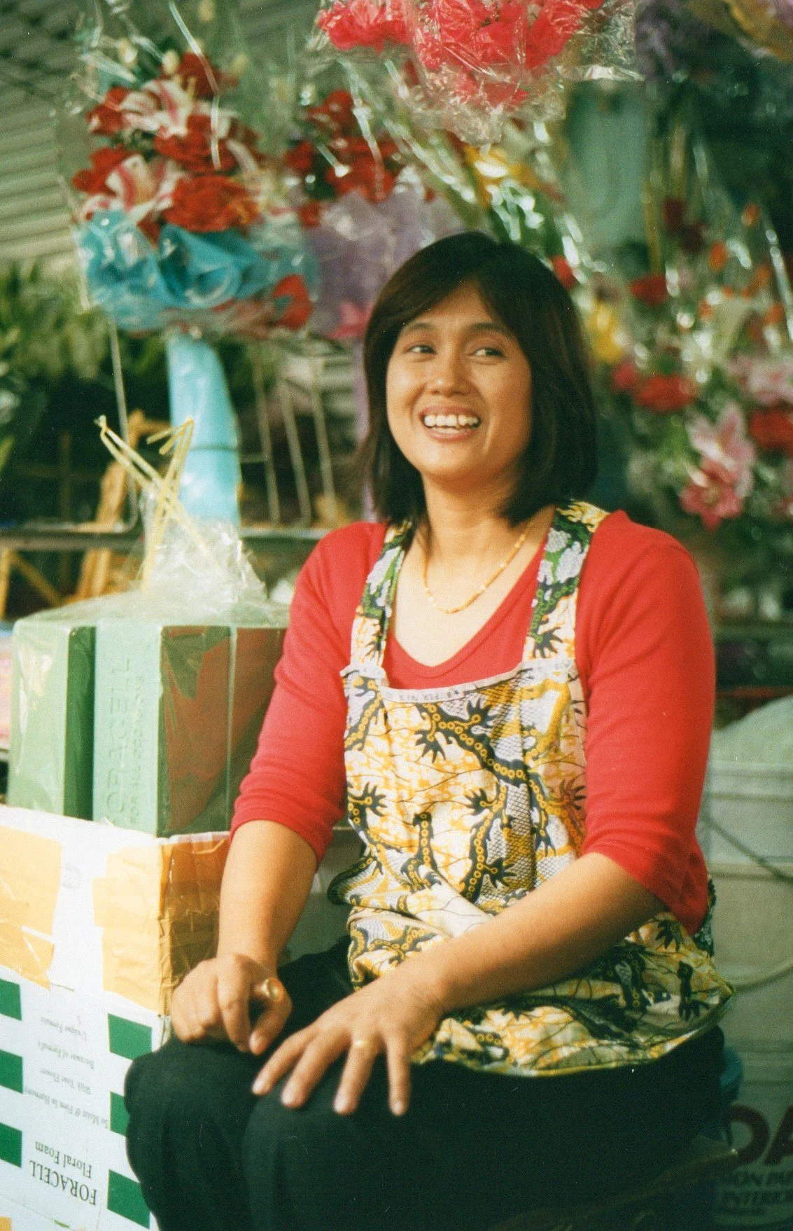 A woman smiling, wearing a red shirt and patterned apron, sitting in front of a colorful display of flowers and gift boxes.
