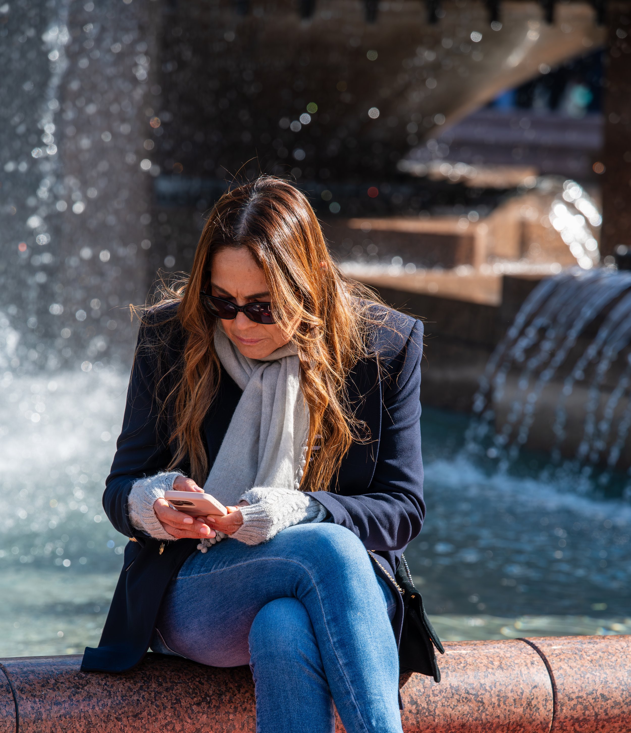Woman with brown hair, sunglasses, navy jacket, scarf, and gloves sitting by water fountain, looking at her smartphone.