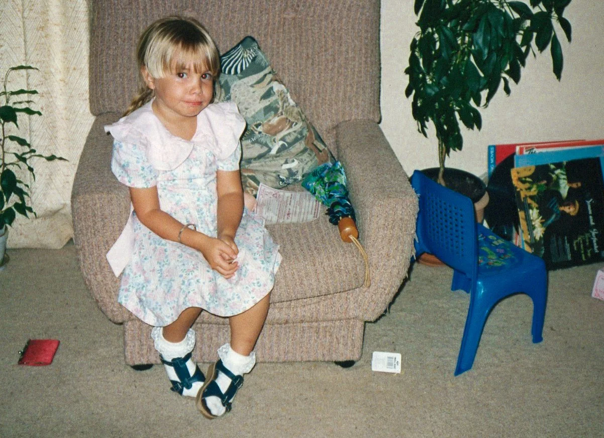 A young girl with blonde hair sitting on a beige armchair in a room with a carpeted floor. She is wearing a floral dress, white socks, and sandals. There is a backpack with a umbrella and some papers on her lap. To the right, there's a small blue plastic chair, a potted plant, and a few books and magazines on the floor.