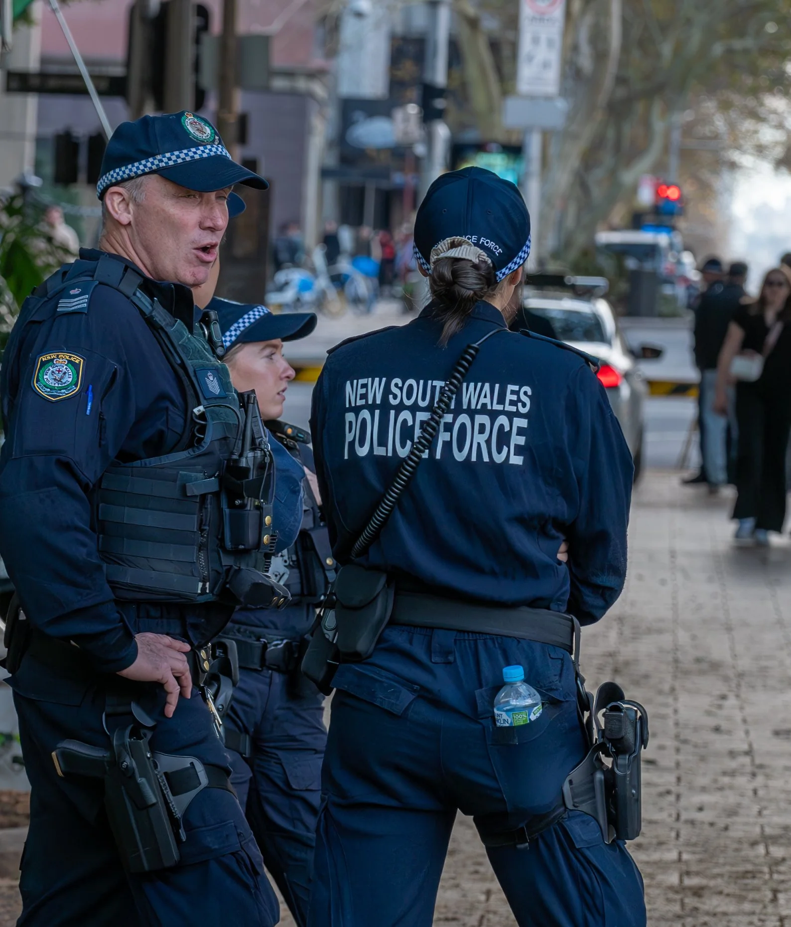 Three police officers from the New South Wales Police Force standing on a sidewalk in an urban area, with a police car and pedestrians in the background.