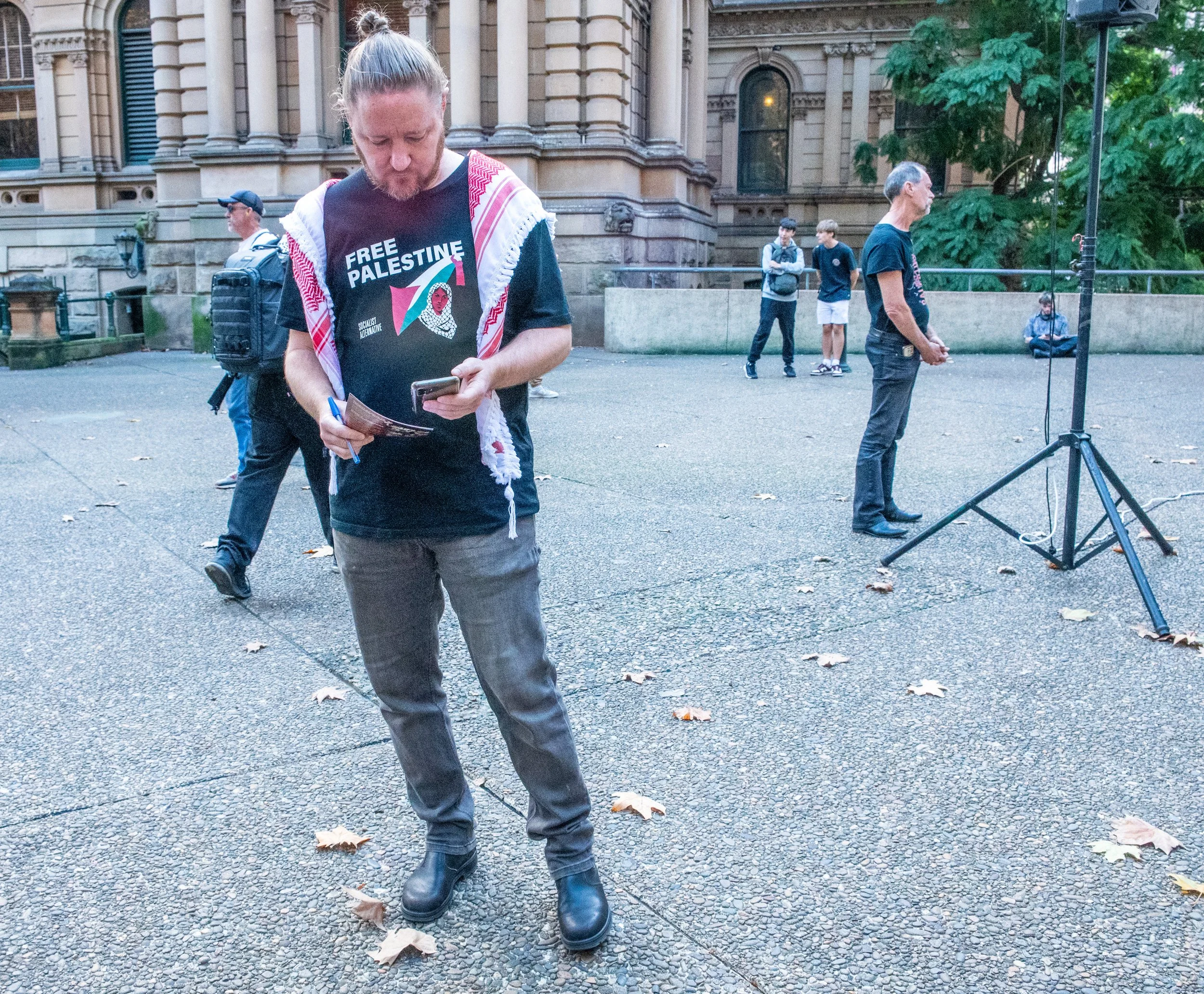 A man wearing a T-shirt with 'Free Palestine' writing and a scarf around his shoulders is standing on a paved area, looking at his phone and holding a flyer. Several other people are in the background, some standing and some sitting, with a historic building and trees in the background.