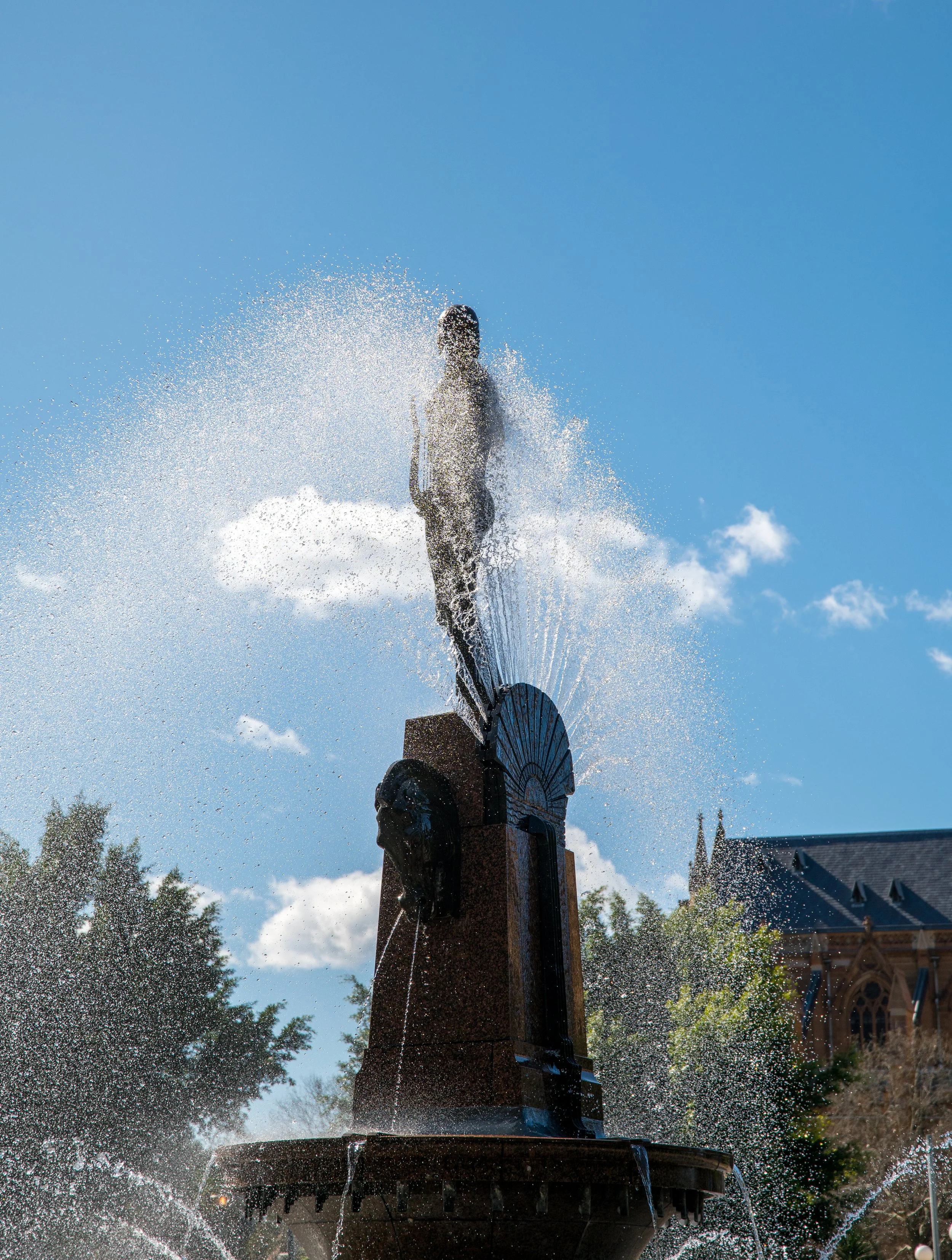 A statue with water sprouting from the head and sides, part of a fountain, against a bright blue sky with a few clouds and trees in the background.
