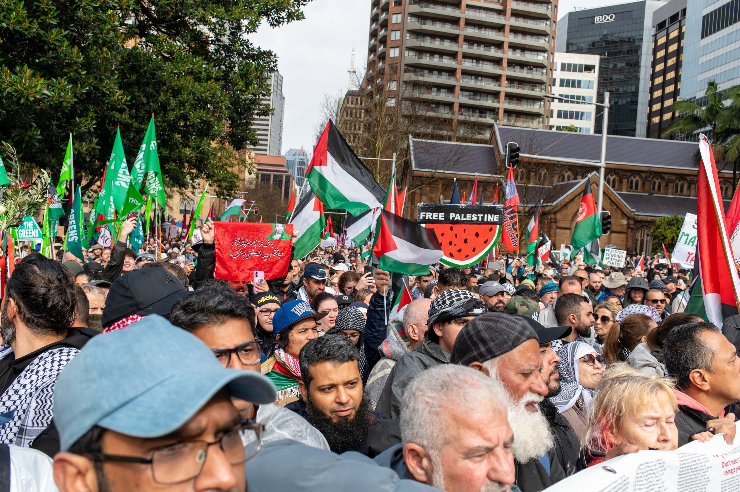 Crowd of protesters holding Palestinian flags and signs during a demonstration in an urban area with tall buildings in the background.