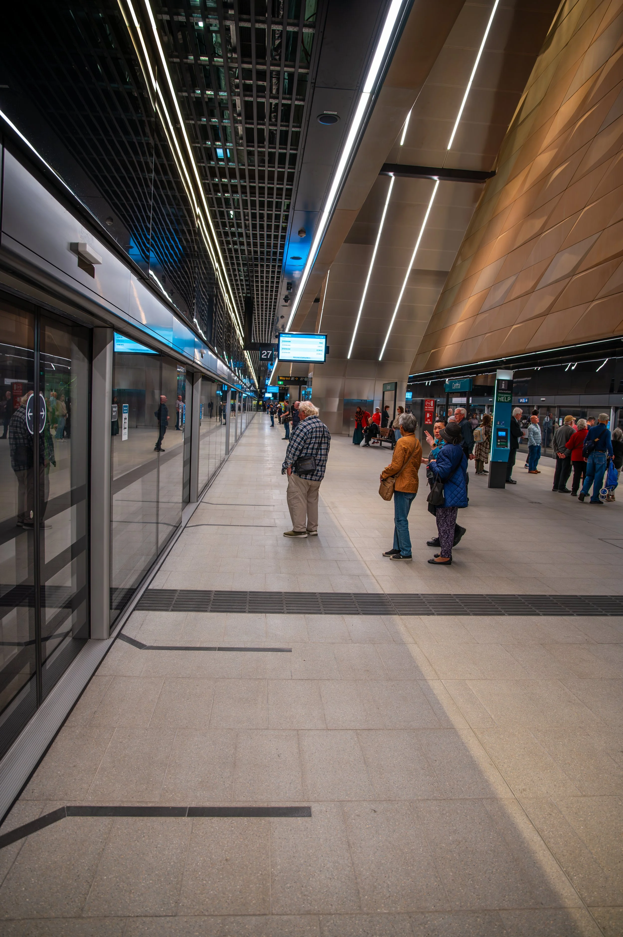 People standing in line at a modern train station platform, with a train on the left side and digital information screens overhead.