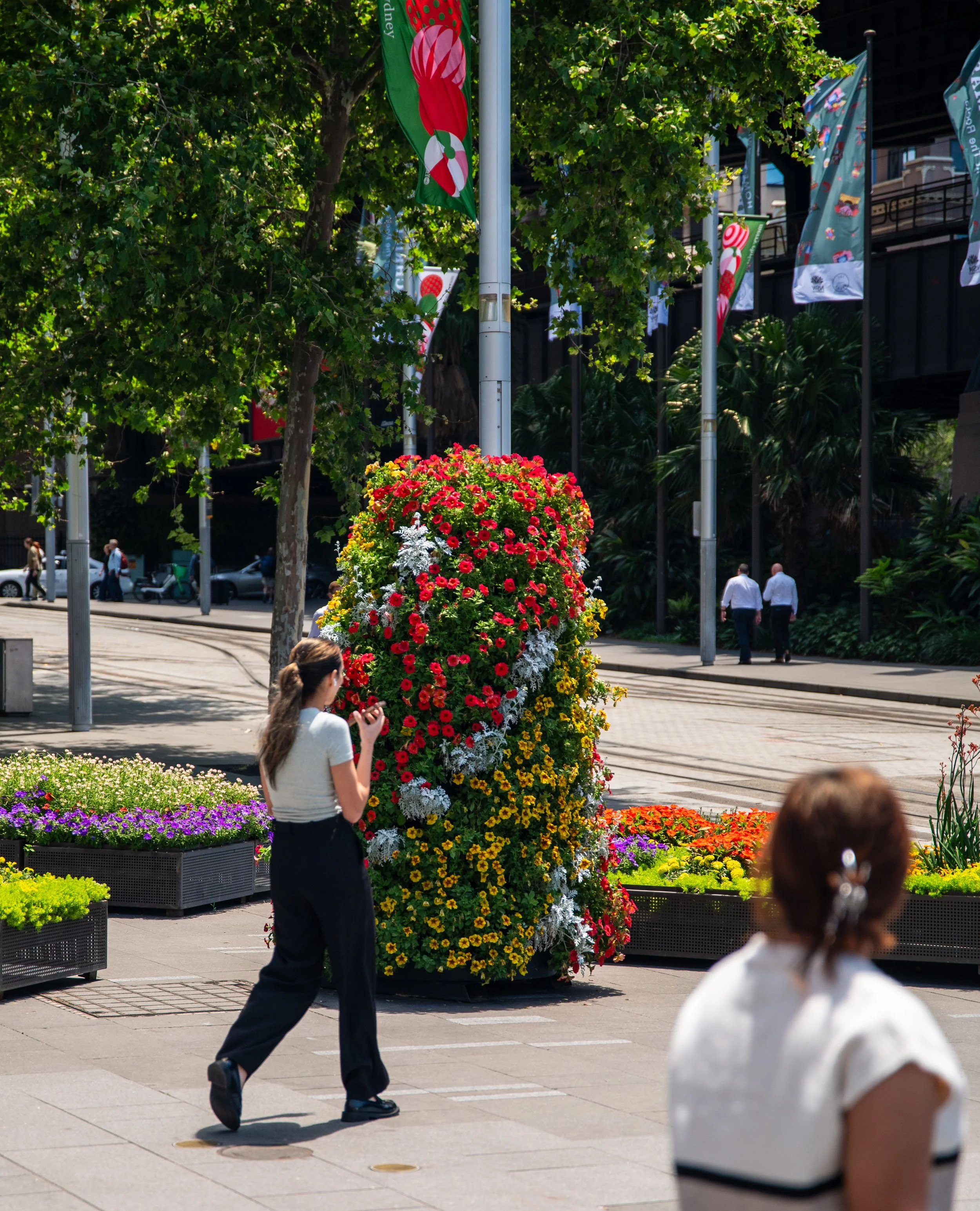 A city street with a large heart-shaped flower arrangement made of red, yellow, white, and purple flowers. Two women are nearby, one with her back to the camera and another facing the flower display, with trees, flag banners, and people walking in the background.