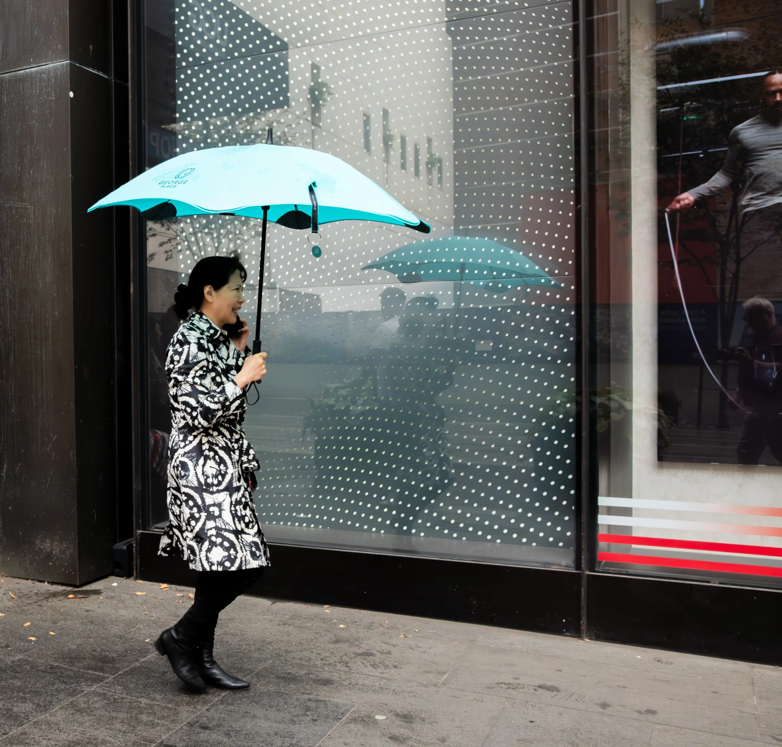 A woman walking on a city sidewalk holds a light blue umbrella, smiling and talking on her cellphone, with a storefront window behind her.