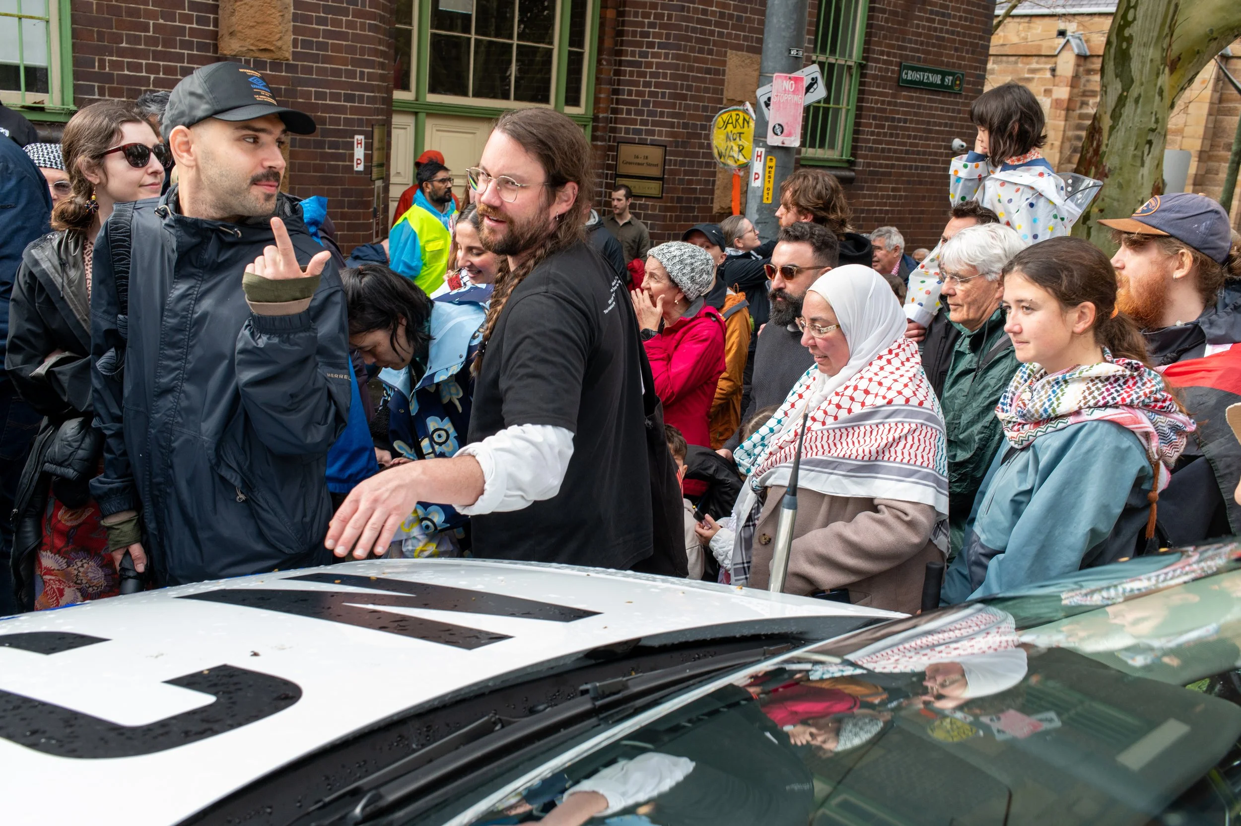 Crowd of diverse people gathered on a city street, some engaging in conversation, with a police vehicle partially visible in the foreground.
