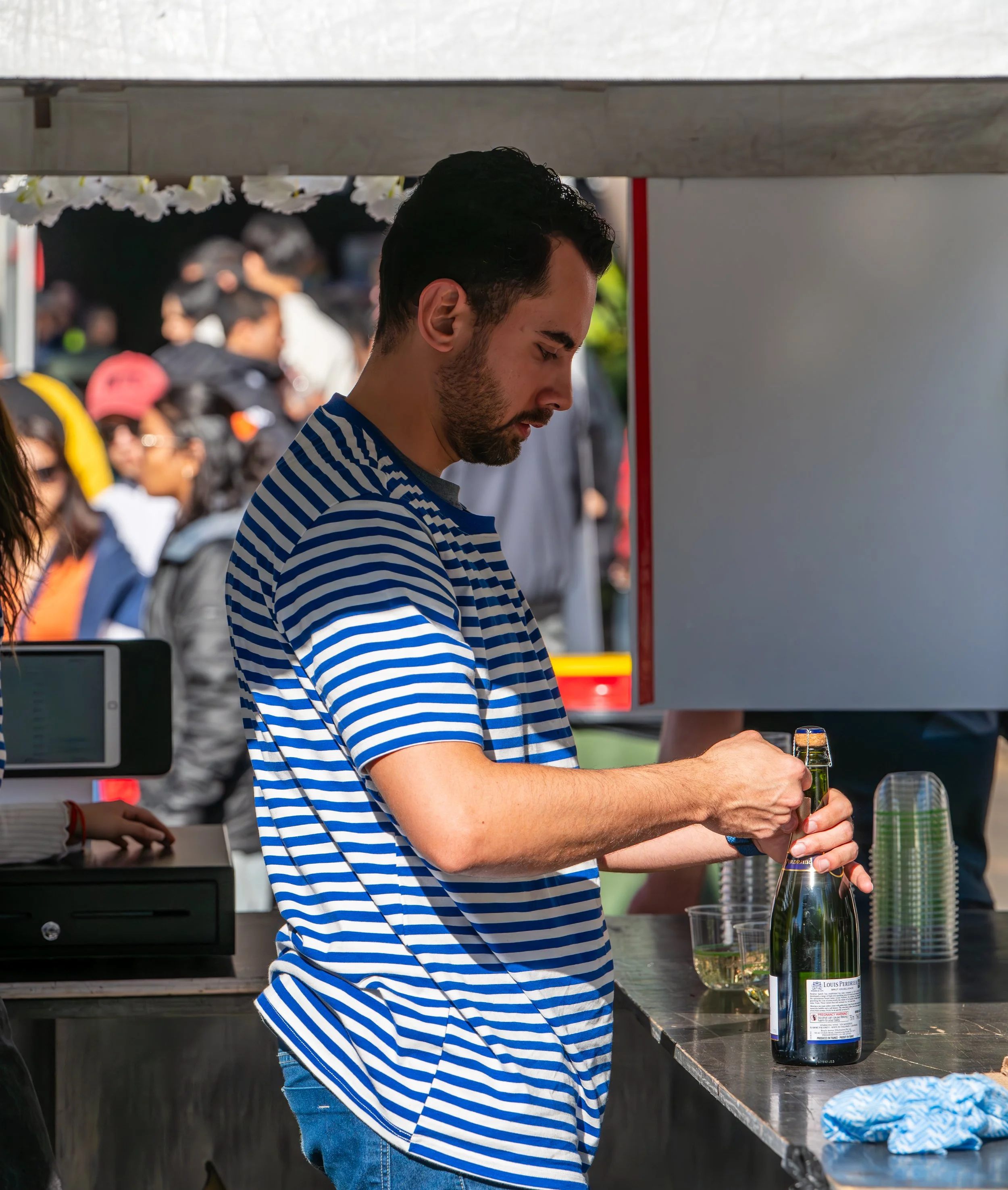 A man with dark curly hair and a beard wearing a blue and white striped t-shirt pouring a drink at an outdoor event, with people in the background.