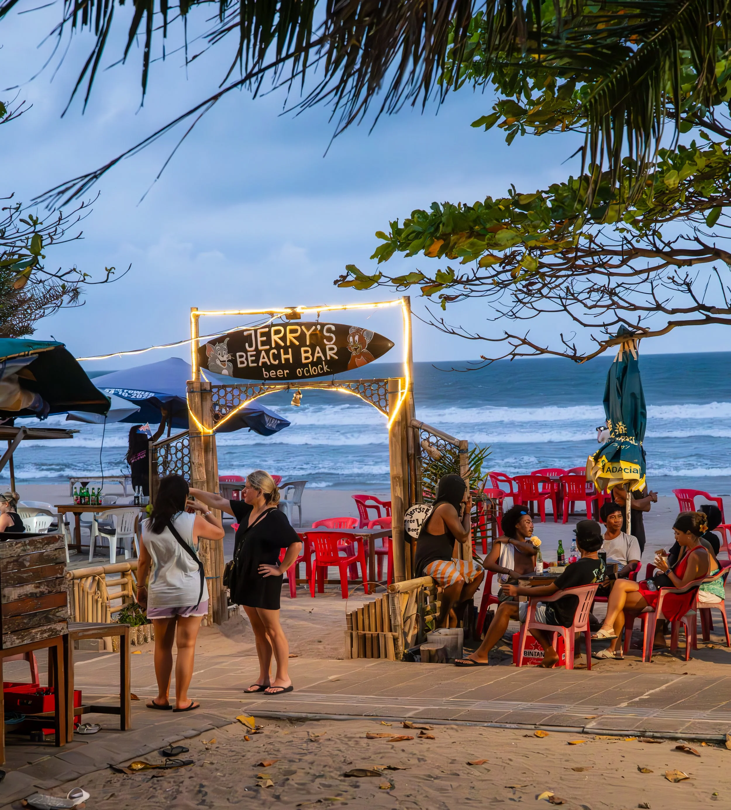 People dining at a beachside outdoor restaurant at sunset with trees and ocean in the background, and a sign reading 'Jerry's Beach Bar' with string lights around the entrance.