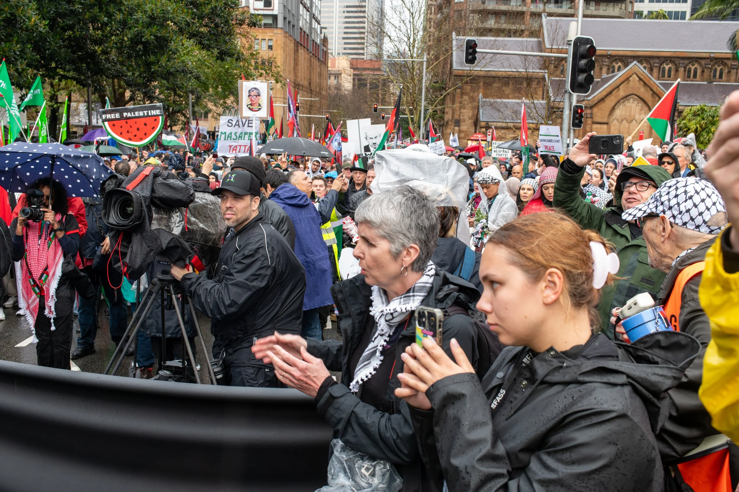 Large crowd of protesters holding signs and flags, some with umbrellas, gathered outdoors in rainy weather during a demonstration.