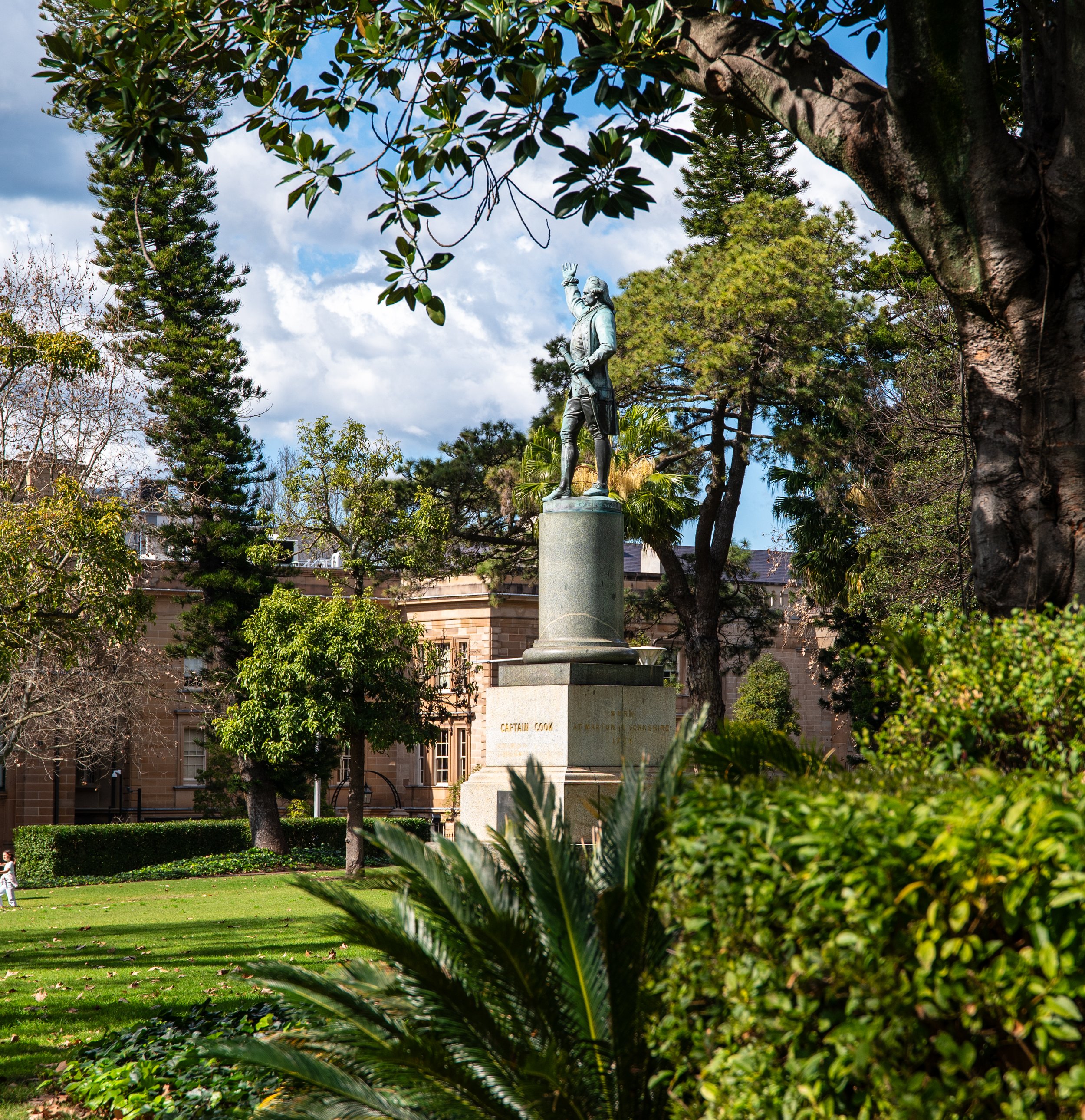 Statue of Captain Cook in a landscaped park with trees, shrubs, and a building in the background under a partly cloudy sky.