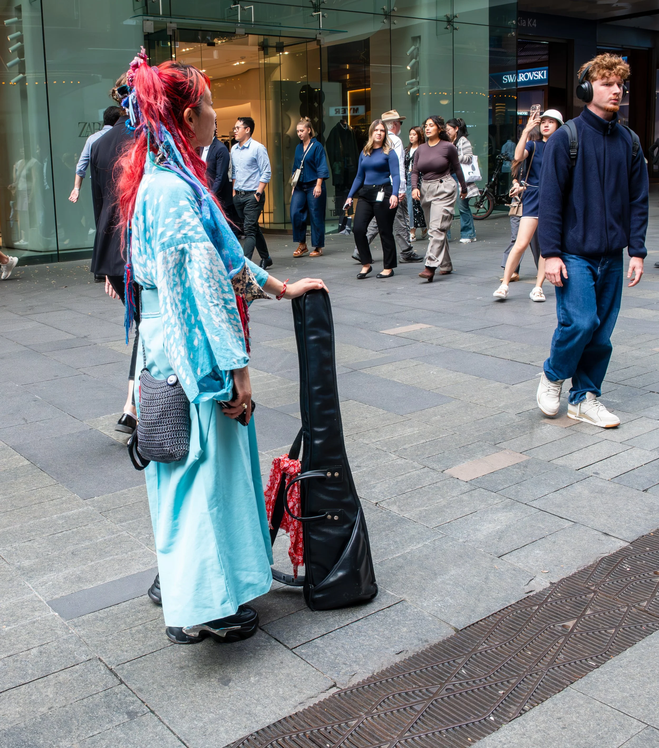 A street performer dressed in a colorful, flowing outfit with bright pink and blue braided hair stands with a guitar case on a busy city sidewalk, surrounded by pedestrians and stores.