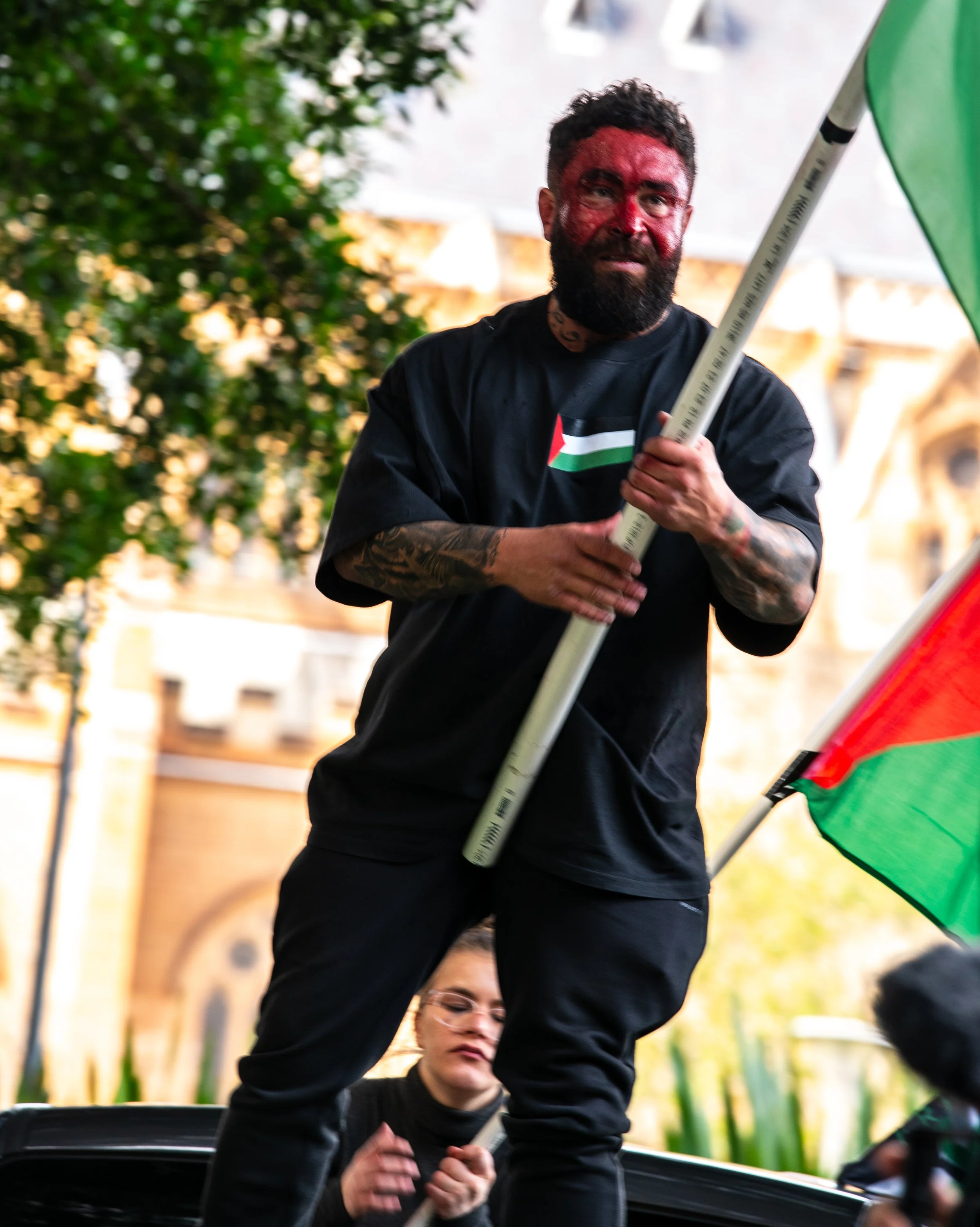 A man with a beard, tattoos, and face paint holding a Palestinian flag at a protest. A woman with glasses is seen below him, holding a flagpole.