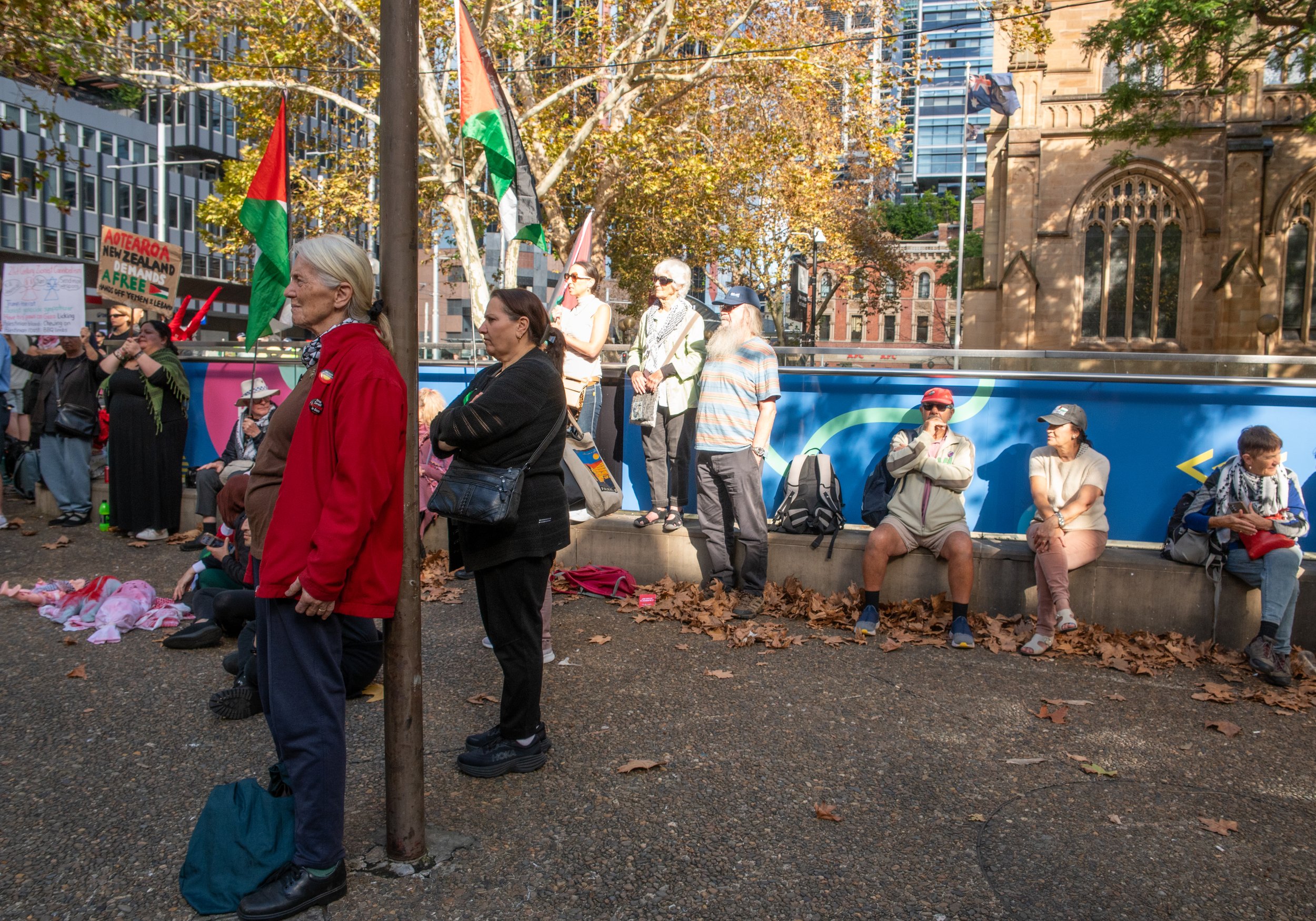 People gathered outdoors for a protest or rally, holding flags and signs; some sit on a concrete ledge, others stand, with a background of trees, buildings, and a blue mural.