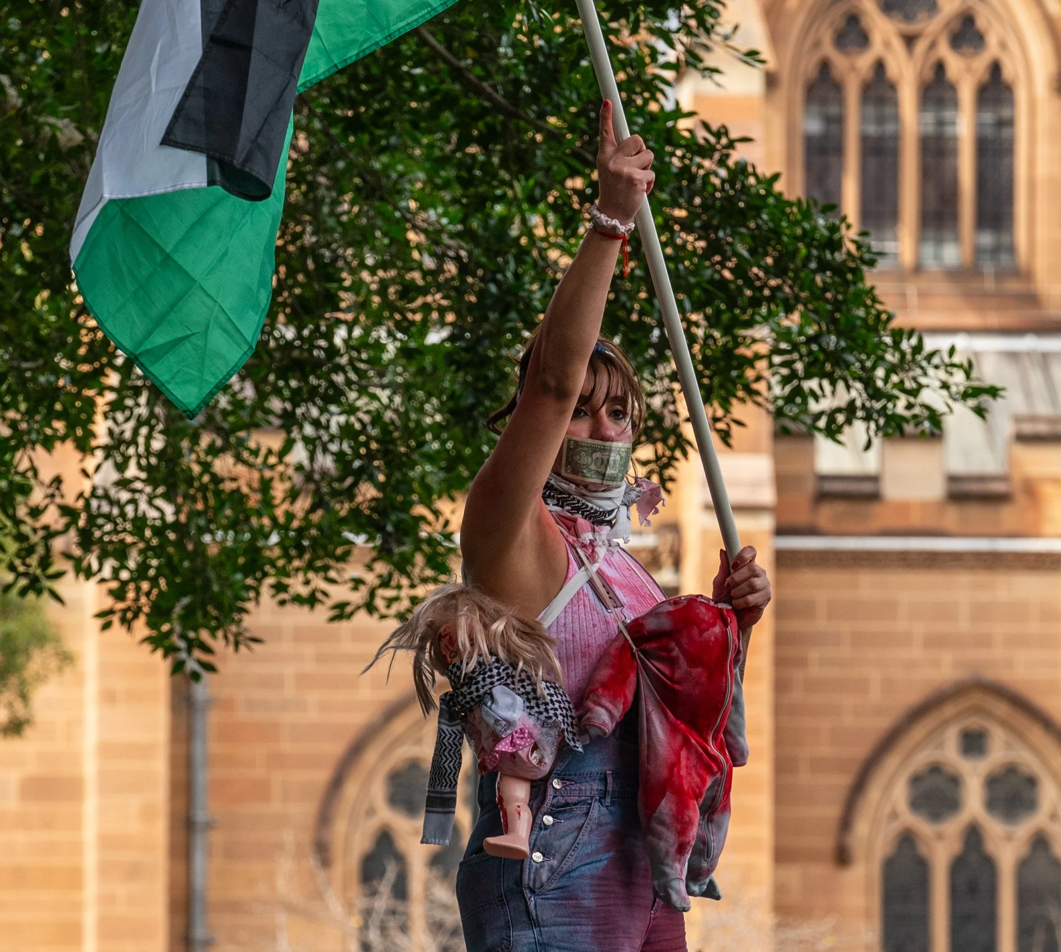 A woman holding a flag with a stuffed doll hanging from her shoulder, wearing a face mask, during a protest or demonstration in front of a historic building.