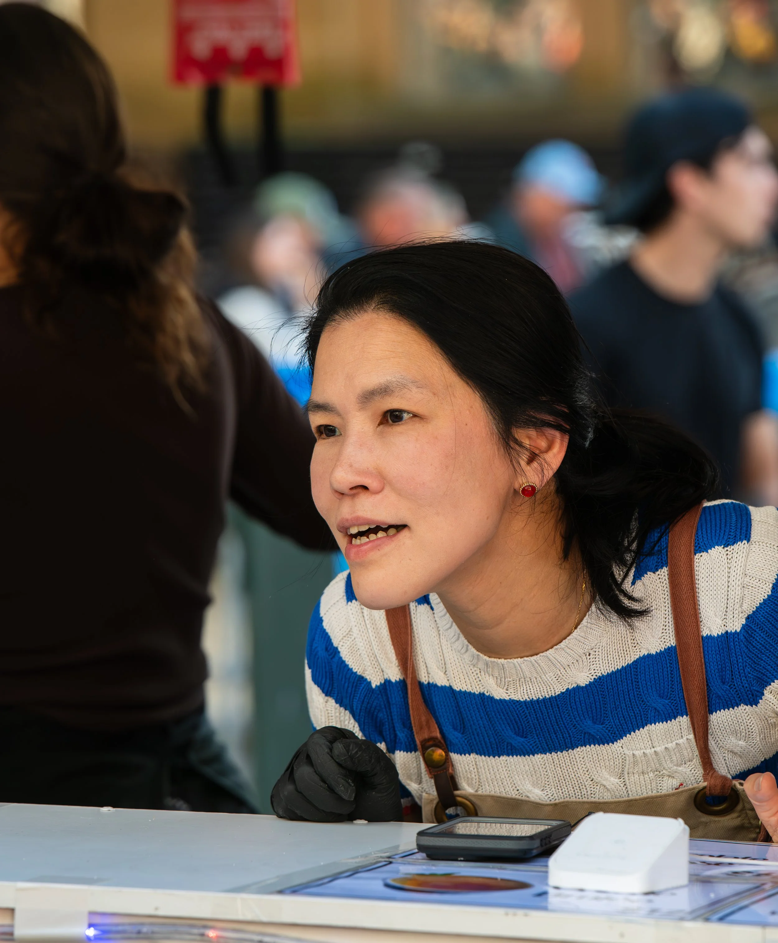 A woman with black hair and red earrings leaning forward, talking to another person at a table during an outdoor event, with a group of people blurred in the background.