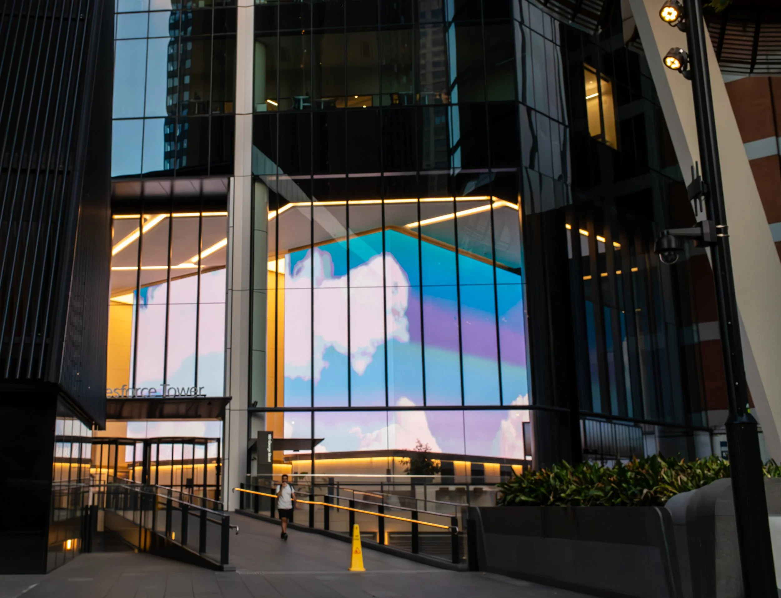 Modern glass building with large digital screen displaying a sky with clouds. A person is walking on a pathway with a yellow caution cone nearby, and the building has the text 'Bellevue Tower'.