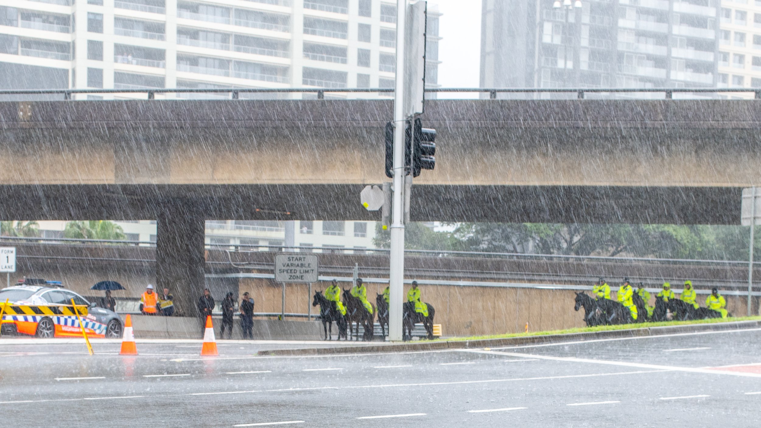 Police officers on horseback and on foot under a bridge during heavy rain, with a police car and traffic cones on the street.