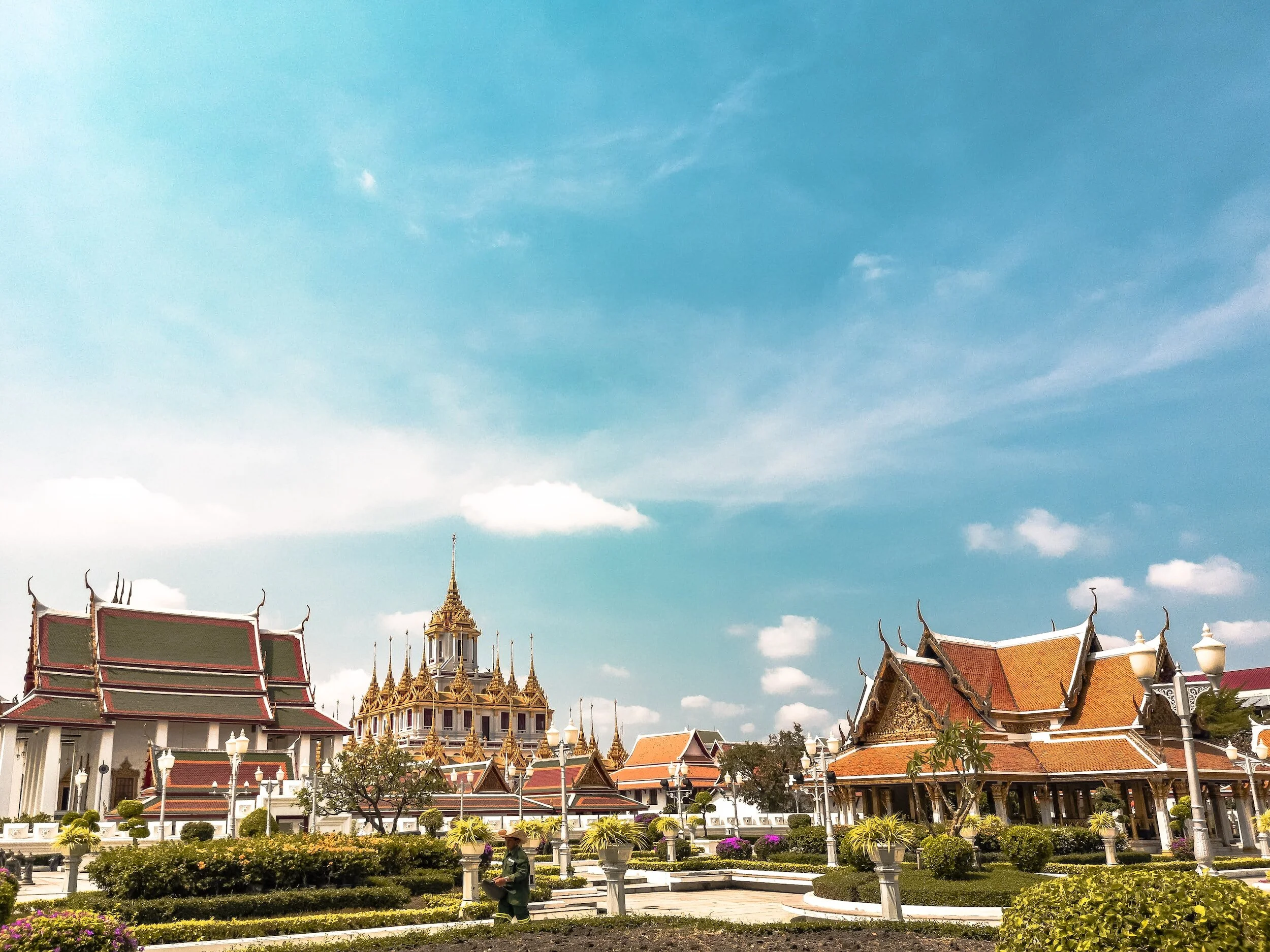Traditional Thai temple with ornate rooftops, garden, and blue sky with clouds.