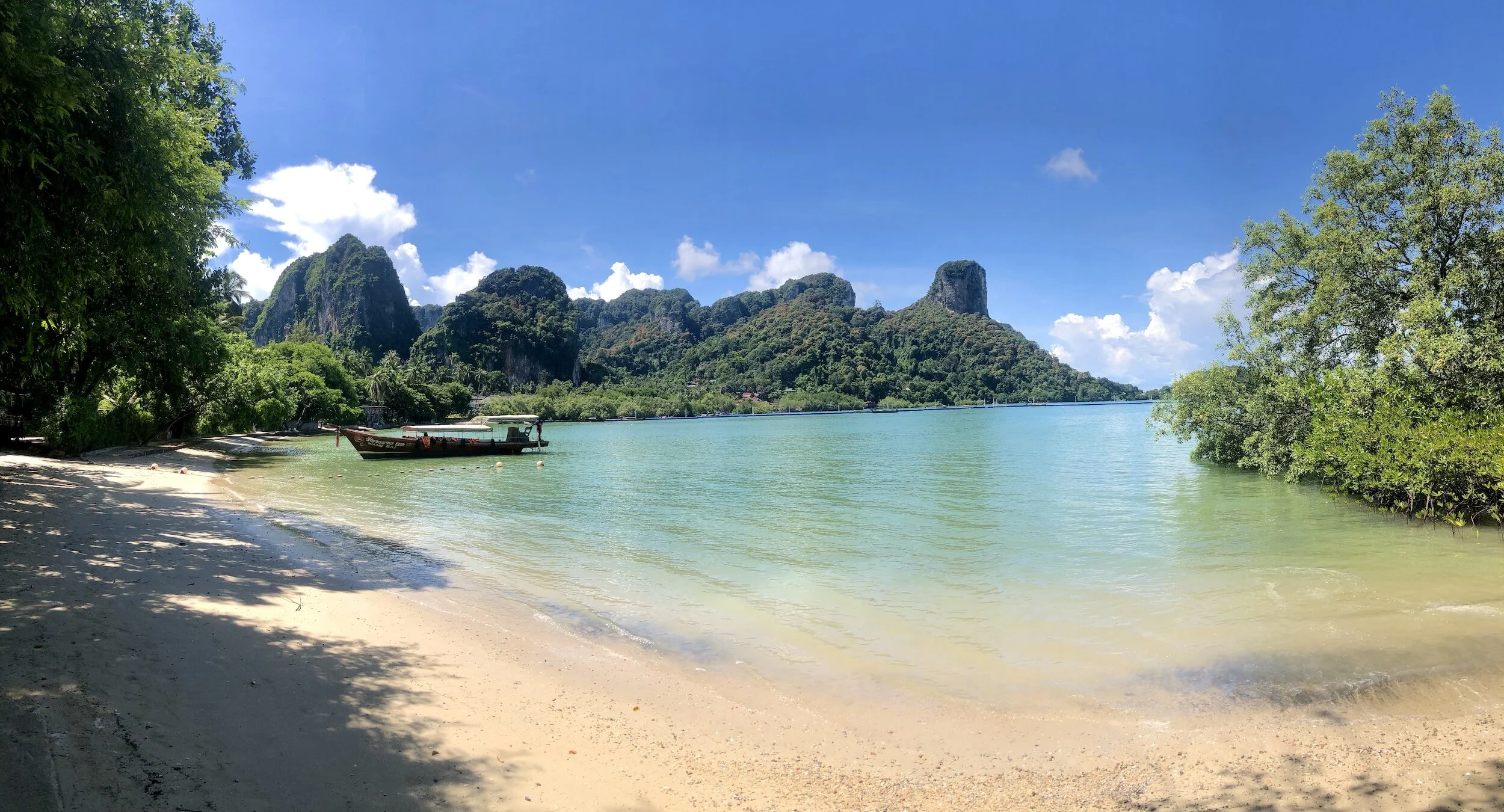 Tropical beach with white sand, clear turquoise water, lush green trees, and dramatic limestone hills under a blue sky with some clouds.