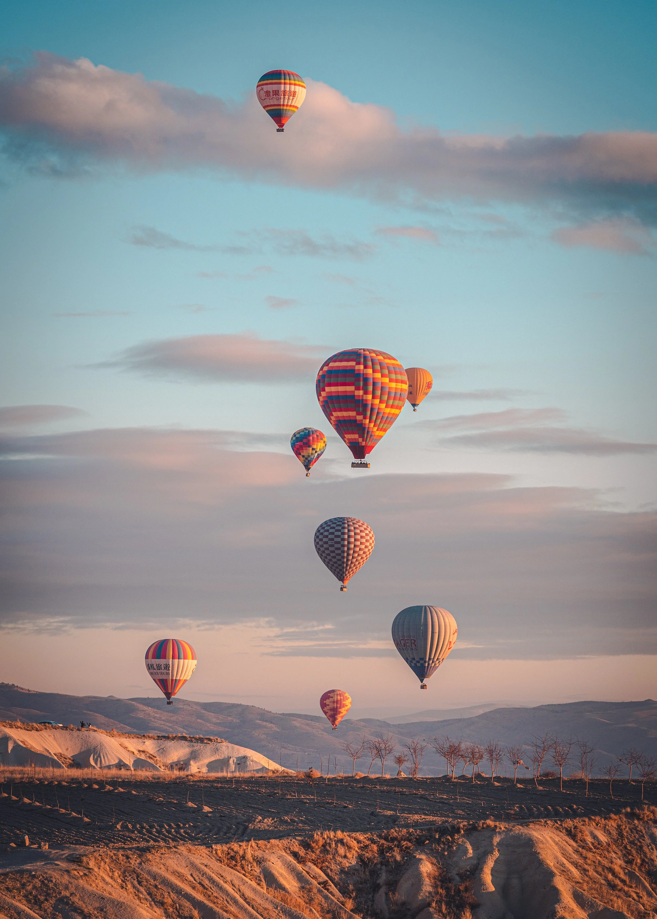 Multiple colorful hot air balloons floating in the sky above a desert landscape with hills and leafless trees.