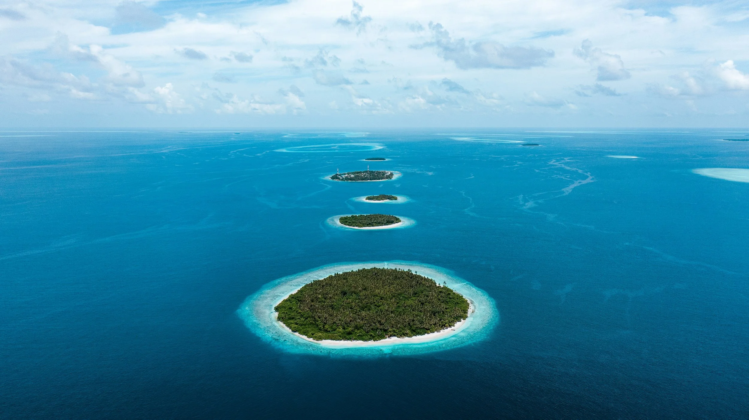 Aerial view of a series of small, green islands with white sandy beaches surrounded by clear blue ocean waters under a partly cloudy sky.